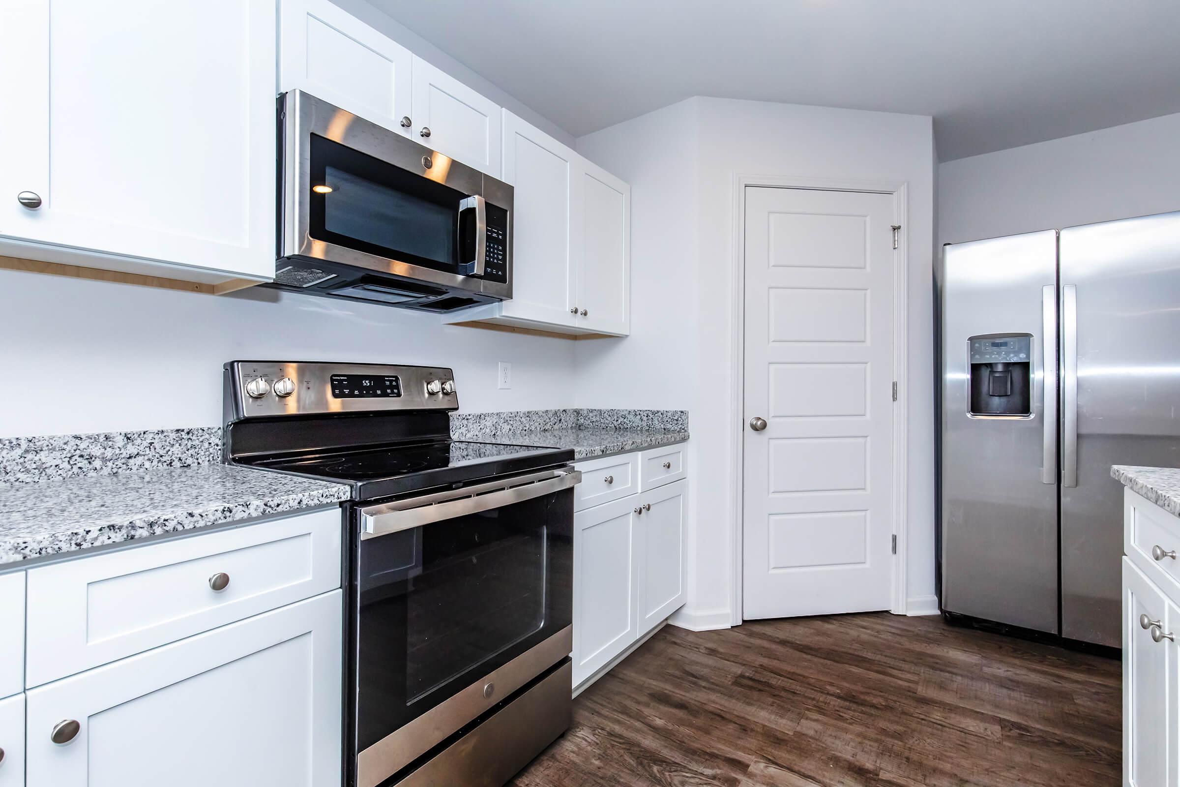 Modern kitchen showcasing stainless steel appliances, including a microwave, oven, and refrigerator. The cabinetry is white, with a granite countertop and wood-style flooring, creating a clean and contemporary look. The layout is open, with a door leading to another area.