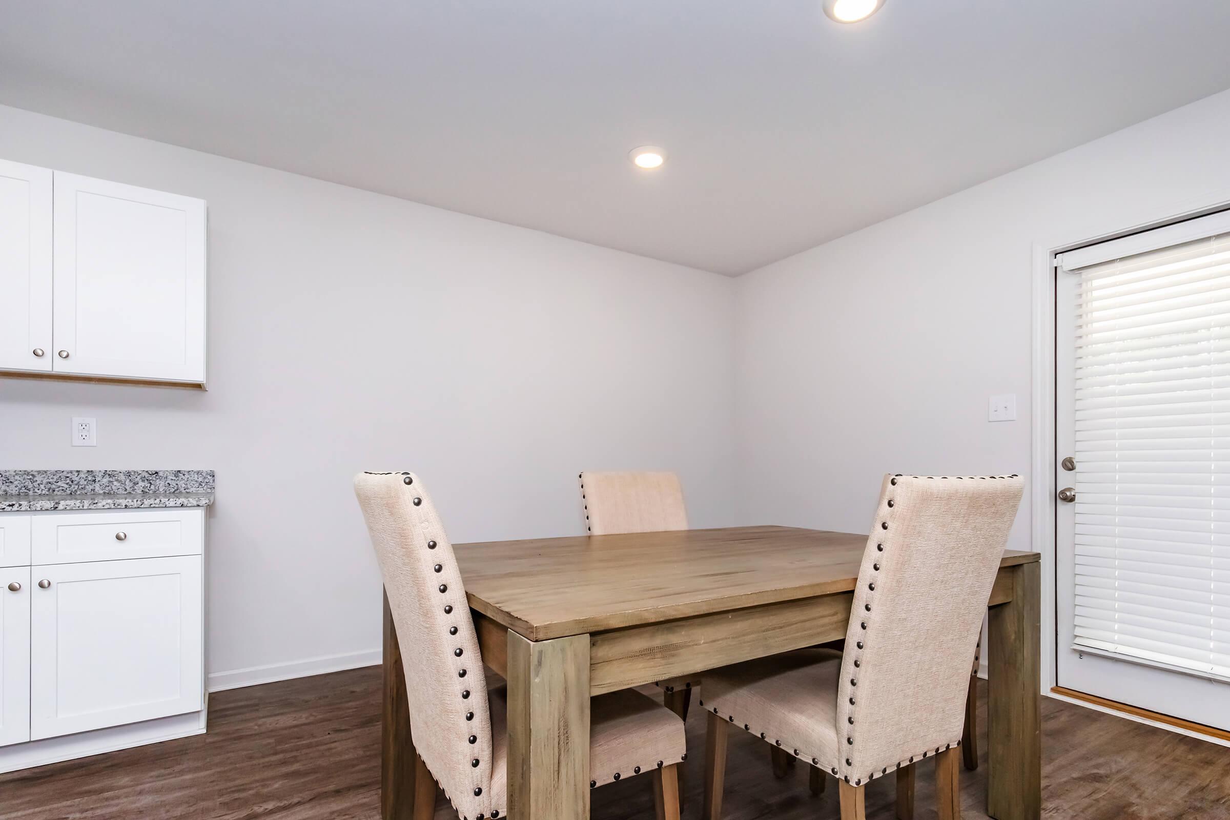A modern dining area featuring a wooden table with four beige upholstered chairs. The walls are painted white, and there is a sliding glass door leading to an outdoor area. White kitchen cabinets are visible in the background along with a granite countertop. The flooring is a warm brown tone.