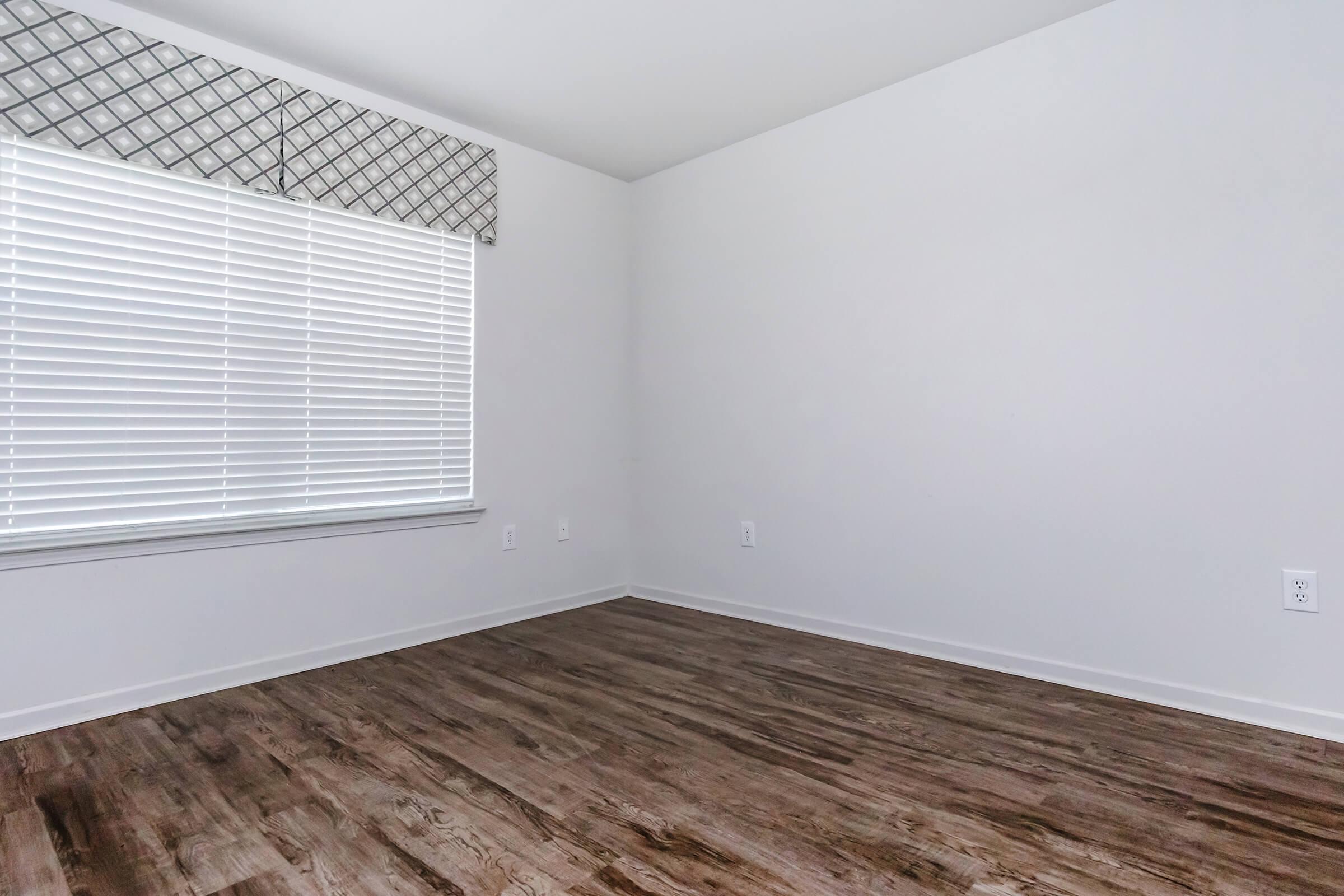 A bright, empty room with a window featuring white blinds. The walls are painted in a neutral color and the floor is made of hardwood, giving a clean and spacious appearance. There is no furniture in the room, highlighting its openness and potential for decoration.
