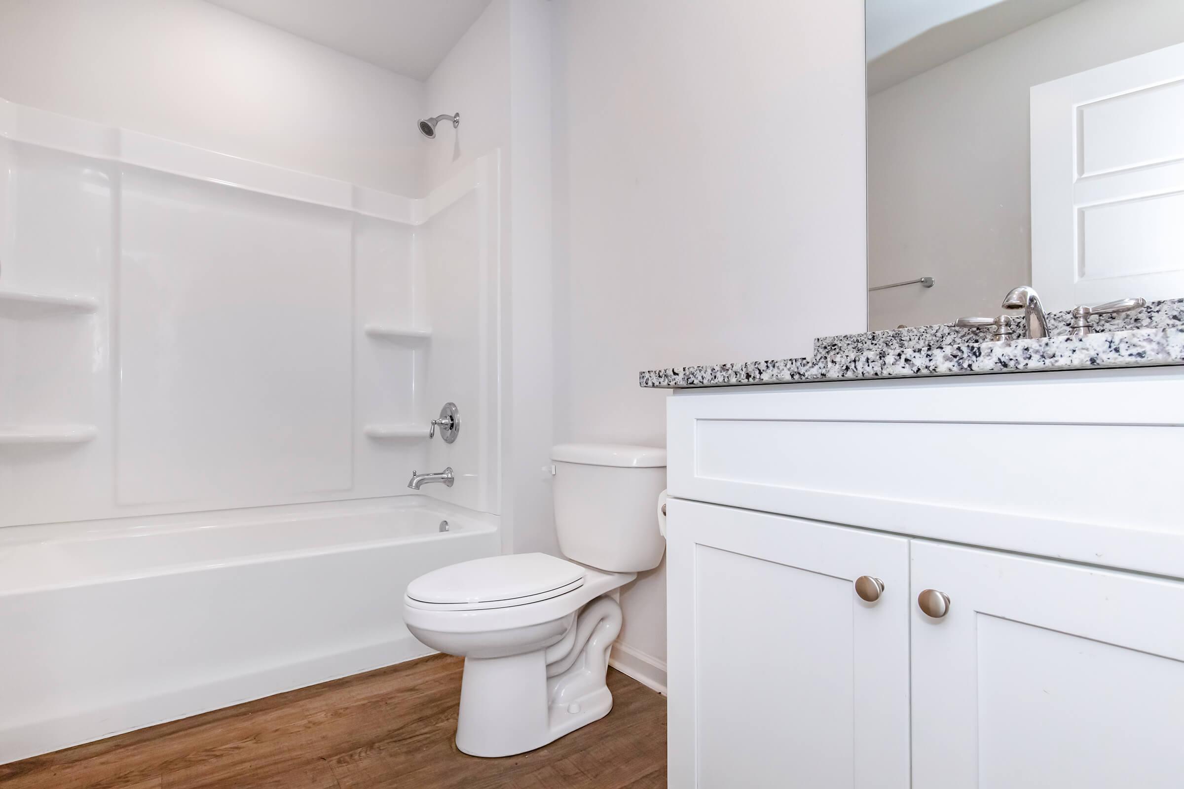 A clean and modern bathroom featuring a white bathtub against a light wall, a white toilet, and a white vanity with a granite countertop. Natural light brightens the space, enhancing the sleek, minimalist design with wooden flooring and a simple cabinet door.