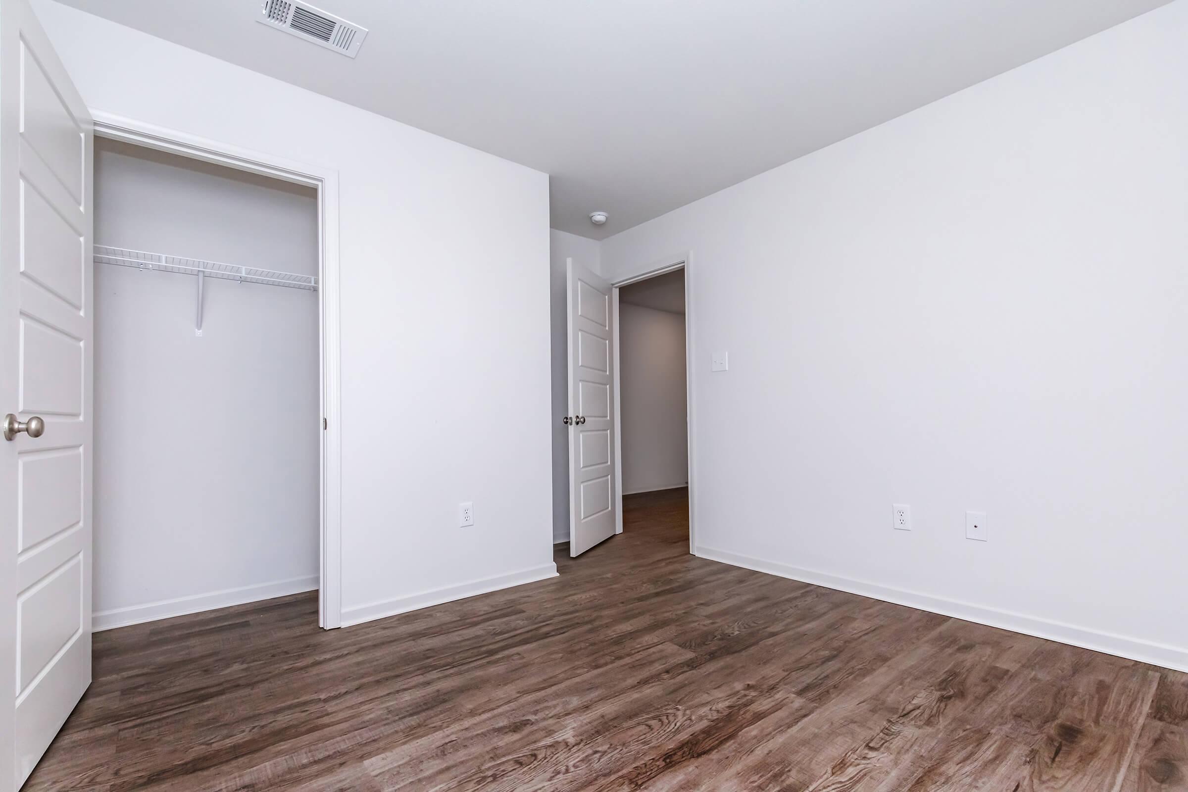 Empty room featuring two doorways, one leading to a closet with shelving and the other to a hallway. The walls are painted white, and the floor is made of wood-like laminate, giving a modern and minimalist aesthetic.
