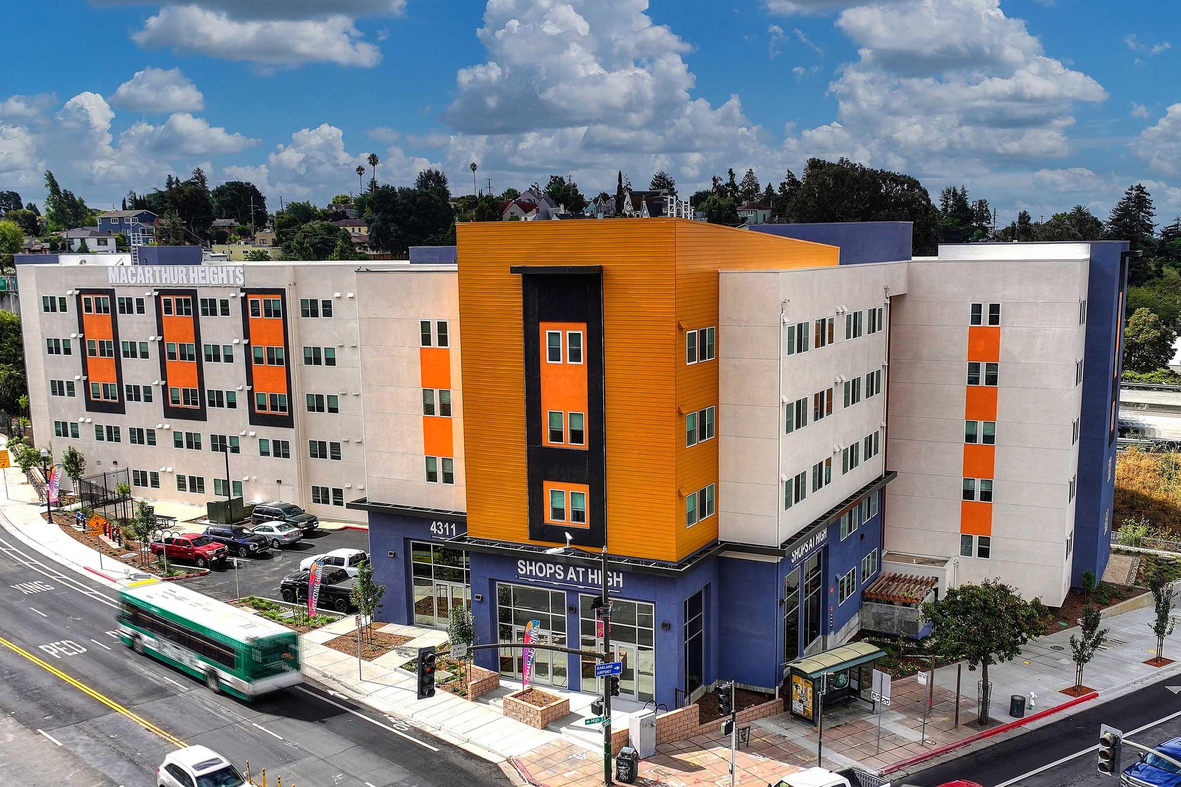A modern multi-story building featuring a mix of orange and light-colored exterior panels. The ground level includes storefronts, and there's a parking area nearby with several cars. In the background, there are trees and a blue sky with scattered clouds. The scene appears to be a vibrant urban area.