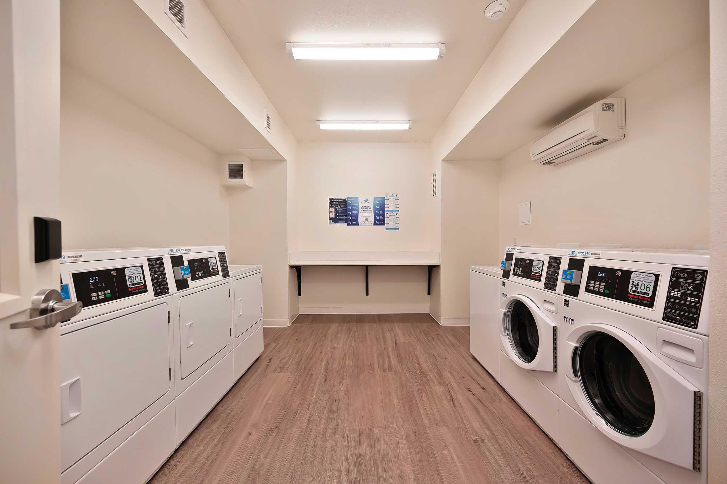 A clean and modern laundry room featuring two sets of white washer and dryer machines arranged side by side. The space has light-colored walls and a wooden floor, with overhead fluorescent lighting. A wall-mounted air conditioner is visible, and there is a folding table against the wall.