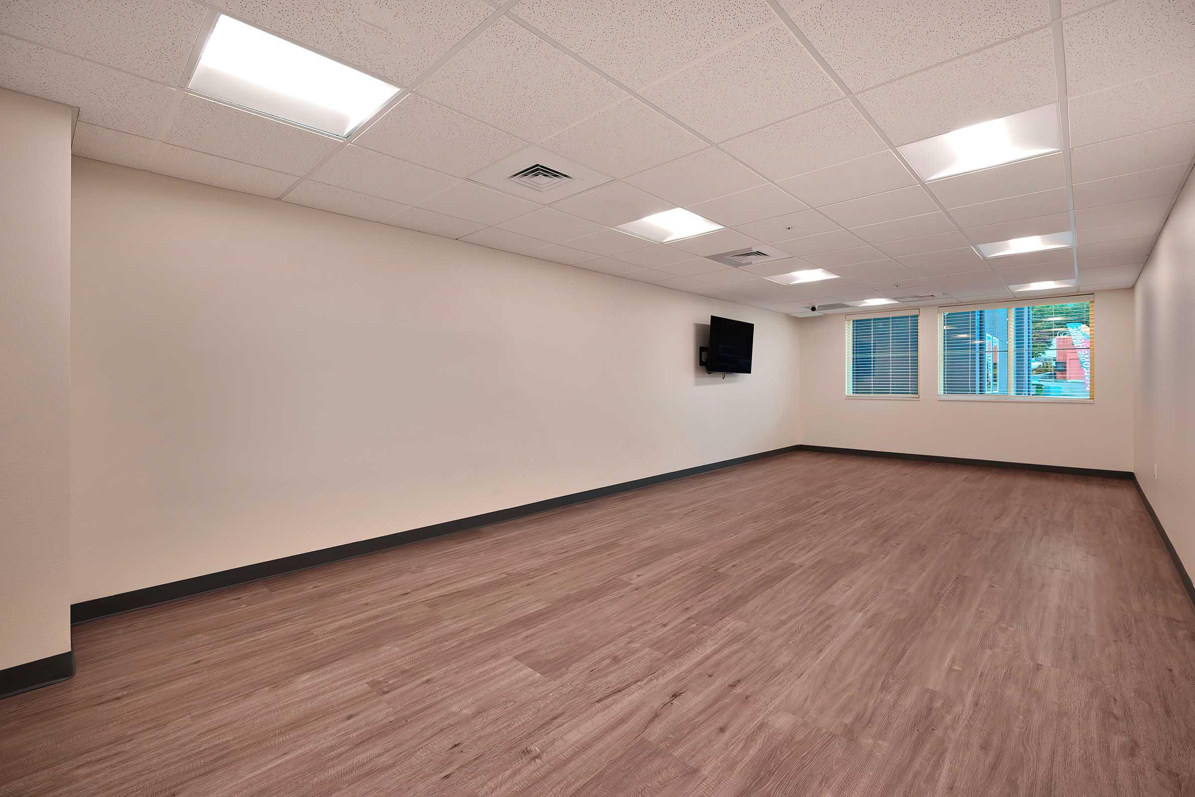 Empty room with light-colored walls and a wood-like floor. The ceiling has fluorescent lights, and a flat-screen TV is mounted on one wall. Windows with blinds allow natural light to enter, but the room appears unoccupied and minimalistic, providing a spacious and clean environment.
