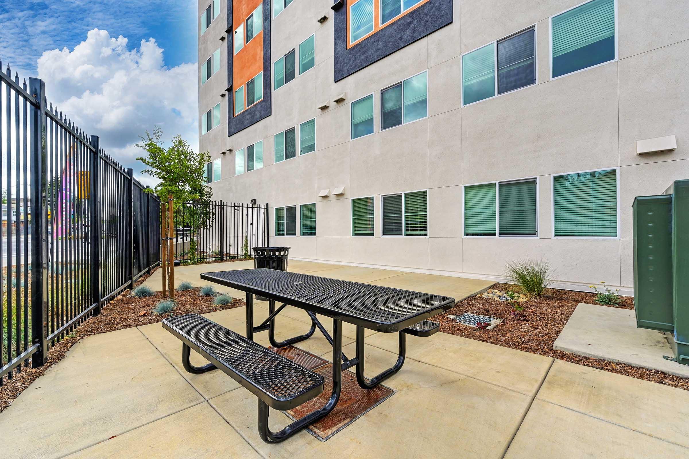 A metal picnic table with benches is situated on a concrete patio in front of a modern building. The building features large windows with green accents and has a fenced area with plants, under a partly cloudy sky.