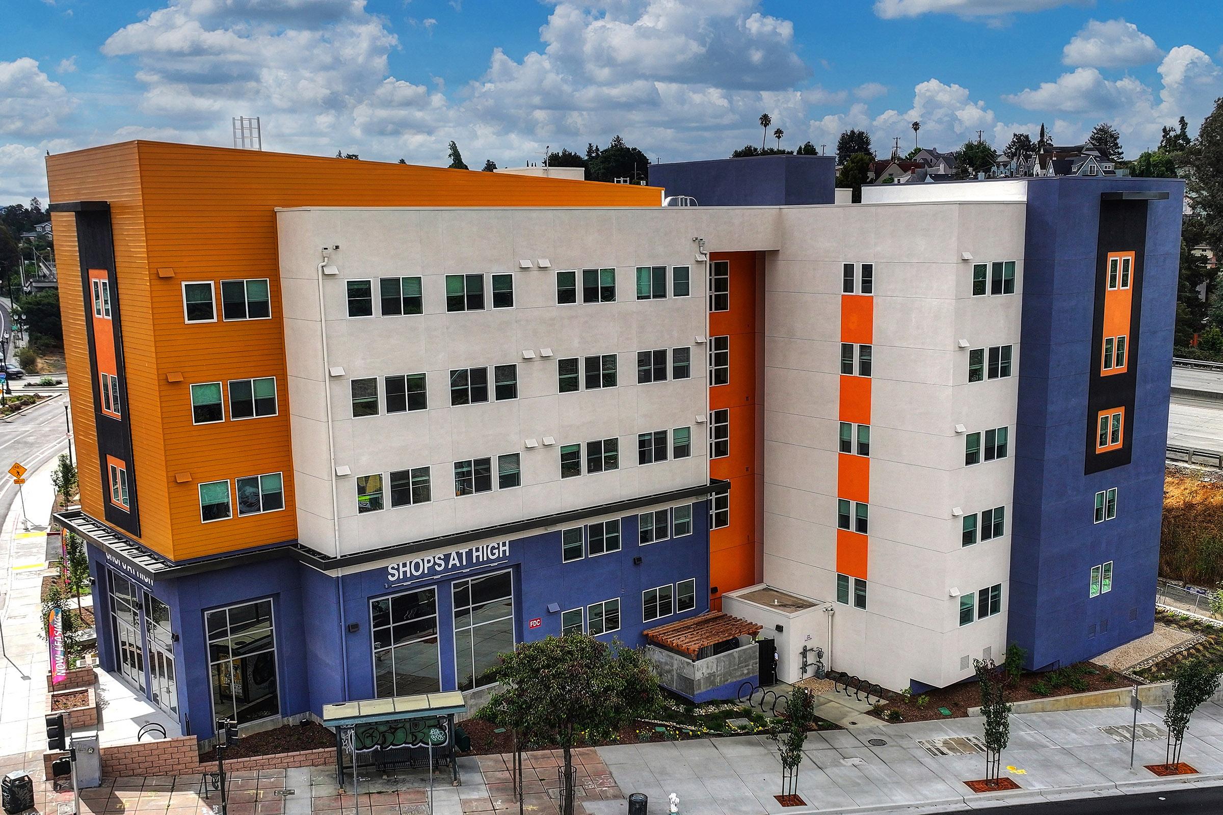 A modern multi-story building with a mix of colors including orange, white, and blue. The facade features large windows and a signage reading "SHOP'S AT HIGH." Surrounding the building are landscaped areas and trees, under a partly cloudy sky. The setting appears urban, likely in a commercial area.