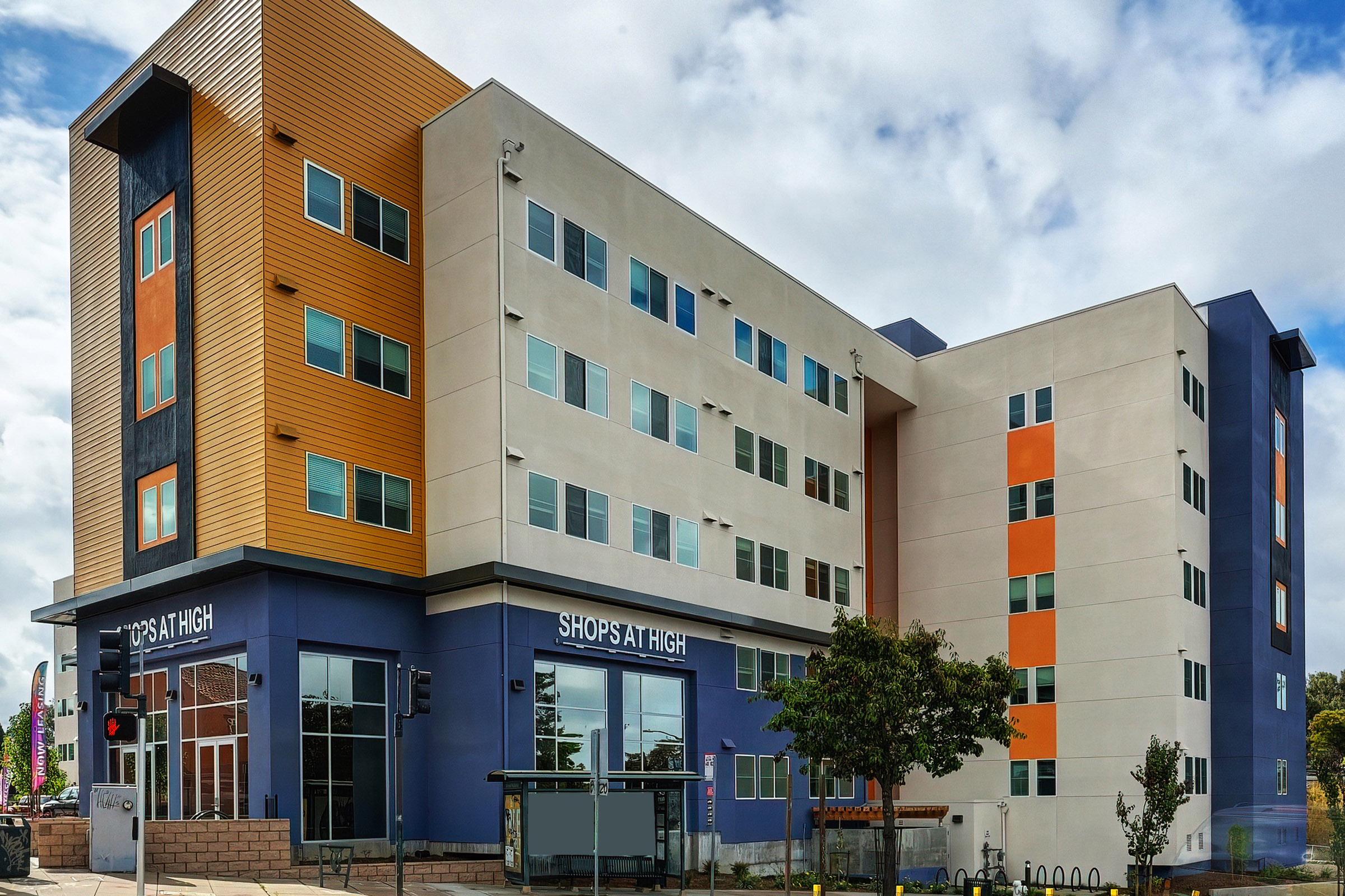 Modern multi-story building with a mix of orange and gray exterior panels. The ground floor features large windows and a business sign "SHOPS AT HIGHT." Surrounding the building are trees and a sidewalk, under a partly cloudy sky.