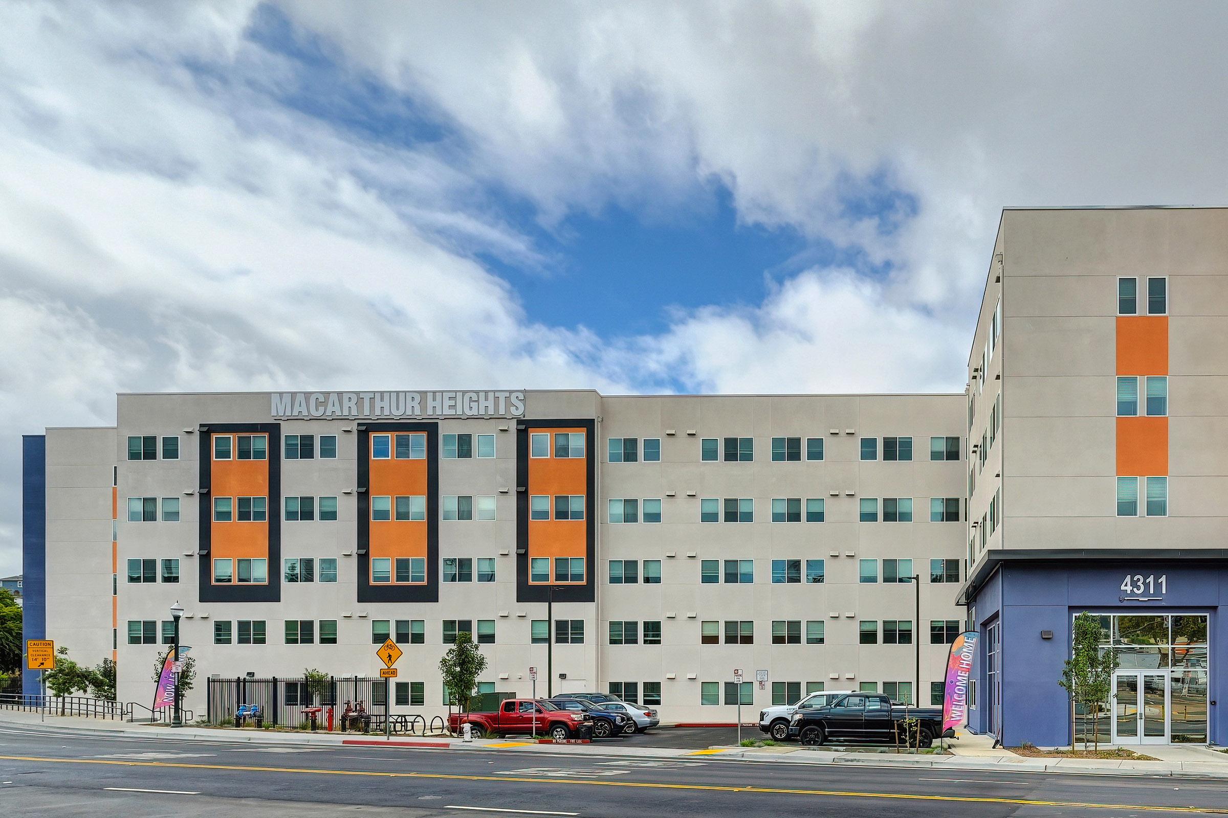 A modern multi-story residential building named "MacArthur Heights" featuring a light-colored facade with orange and blue accents. The building has large windows and is situated on a busy street with parked cars and pedestrian signage visible in the foreground. The sky is partly cloudy.