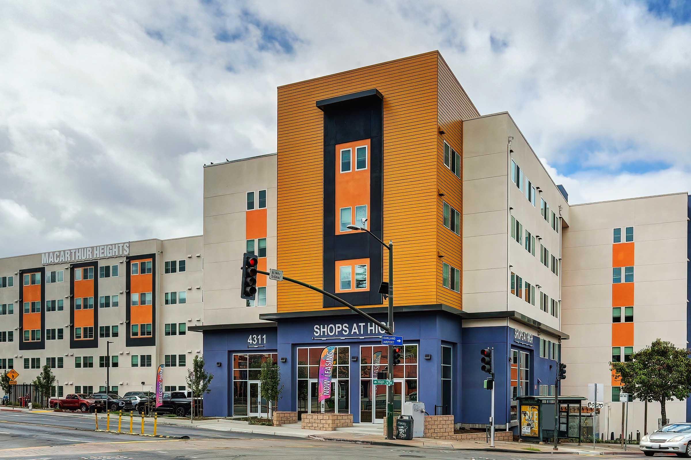 A modern multi-story building with a prominent orange and blue facade, labeled "SHOPS AT," located at the corner of a street. The structure is flanked by additional multi-colored buildings. Traffic lights and street signs are visible, indicating a busy urban area. The sky is partly cloudy.