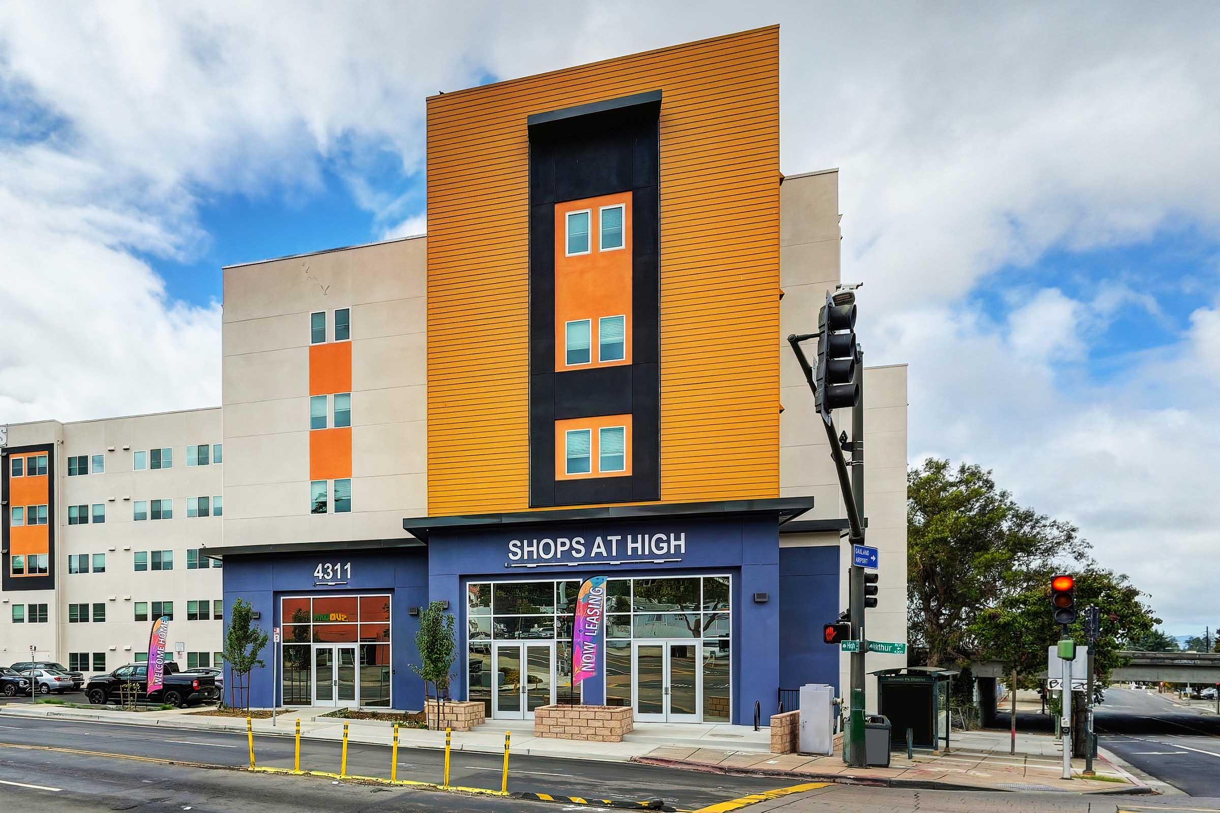 A modern commercial building with vibrant orange and black accents. The storefront features large windows with "SHOPS AT HIGH" displayed prominently. Flanking the entrance are small trees and colorful banners. A traffic signal stands nearby, and the scene is set against a cloudy sky.