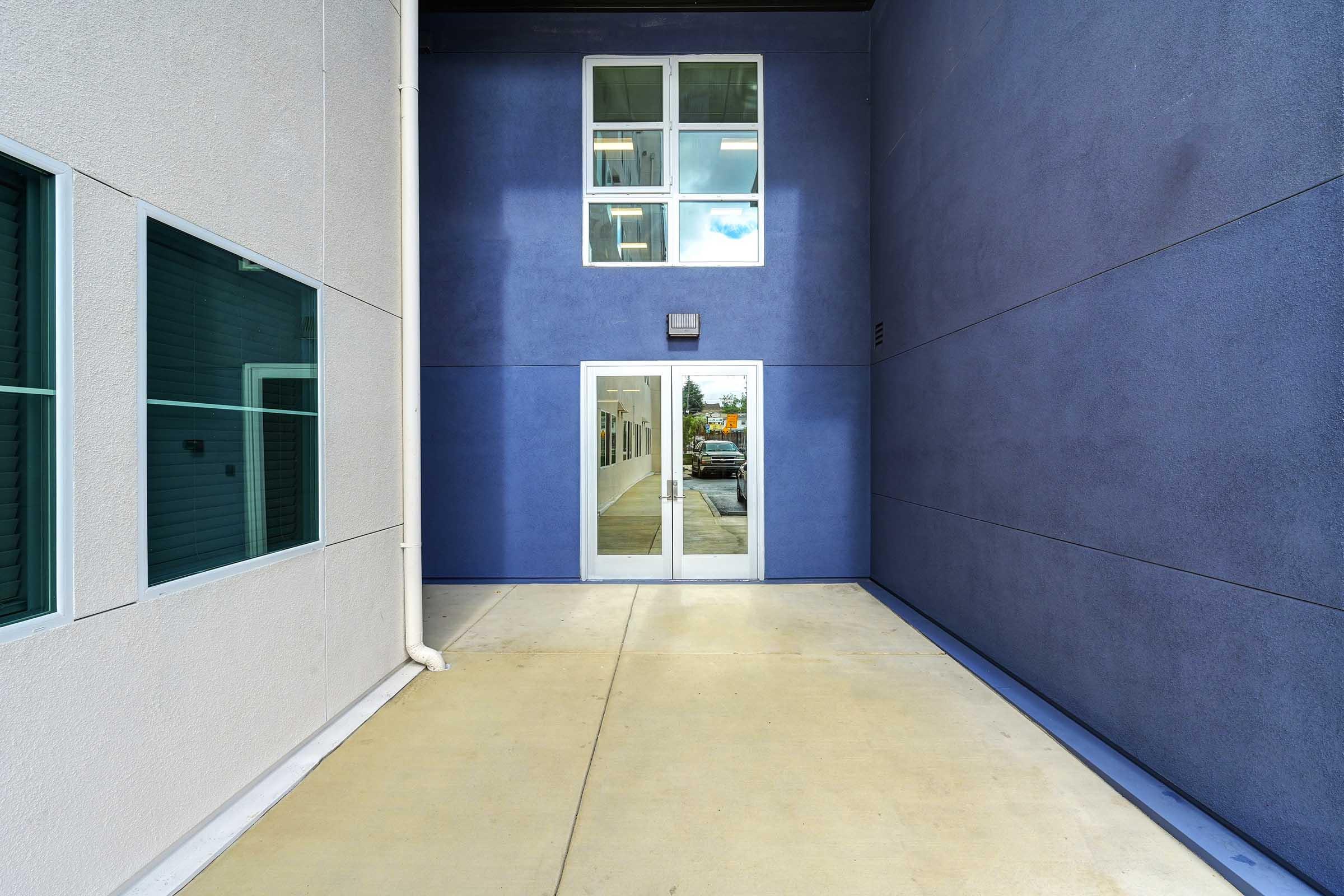 A narrow pathway between two buildings featuring a smooth concrete surface. One side has a light-colored wall with several windows, while the other side displays a dark blue wall. A glass door is centrally located at the end of the pathway, leading to a well-lit interior space.