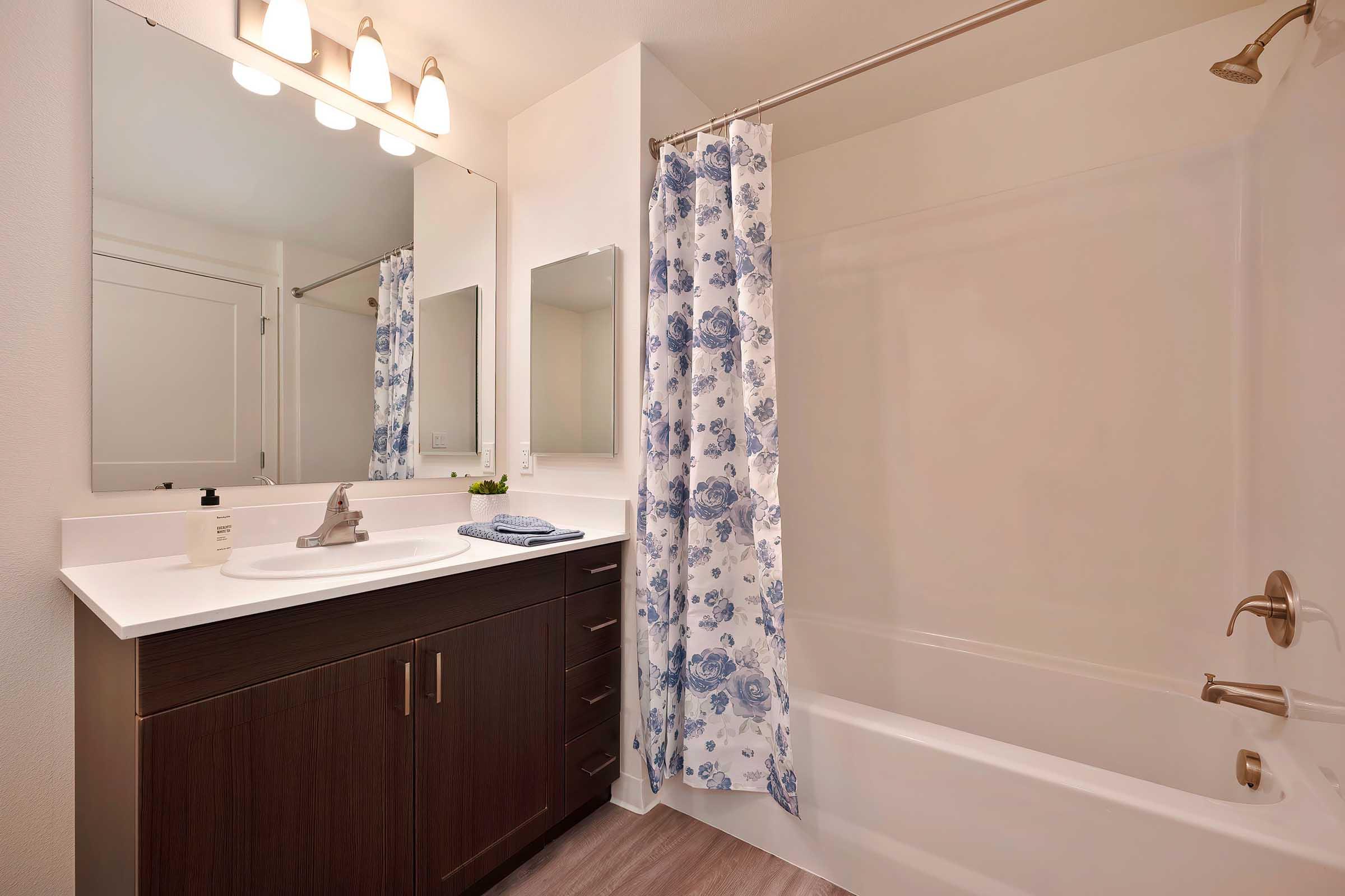 A modern bathroom featuring a bathtub with a shower curtain adorned with blue flowers, a white vanity with a sink, two mirrors above the vanity, and soft lighting provided by three light fixtures above. The floor is wood-like, creating a warm atmosphere.