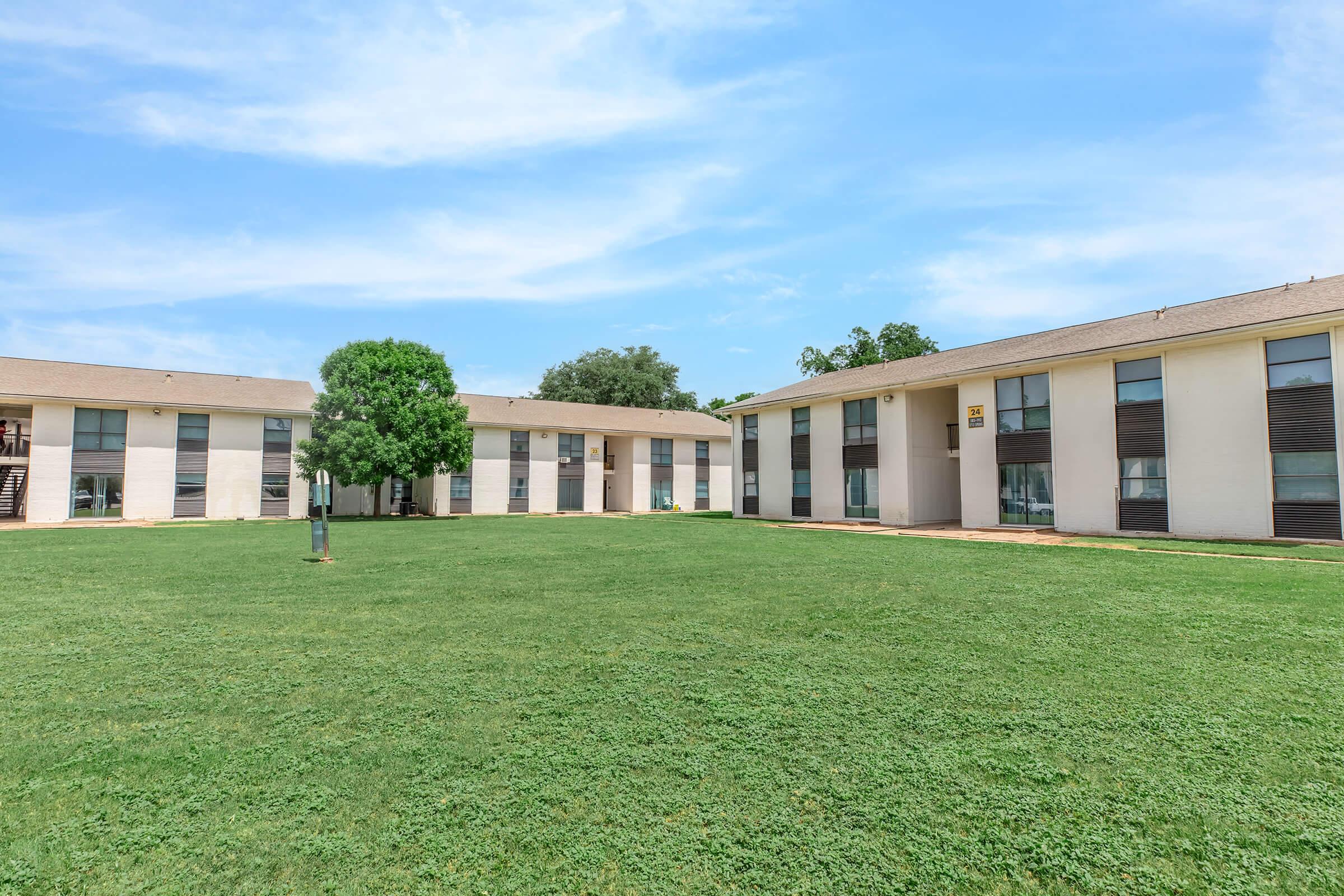 A grassy courtyard surrounded by two residential buildings with large windows. The sky is clear and blue, creating a bright atmosphere. A tree is situated in the middle of the lawn, adding a touch of greenery to the setting. The overall scene conveys a peaceful and inviting environment.