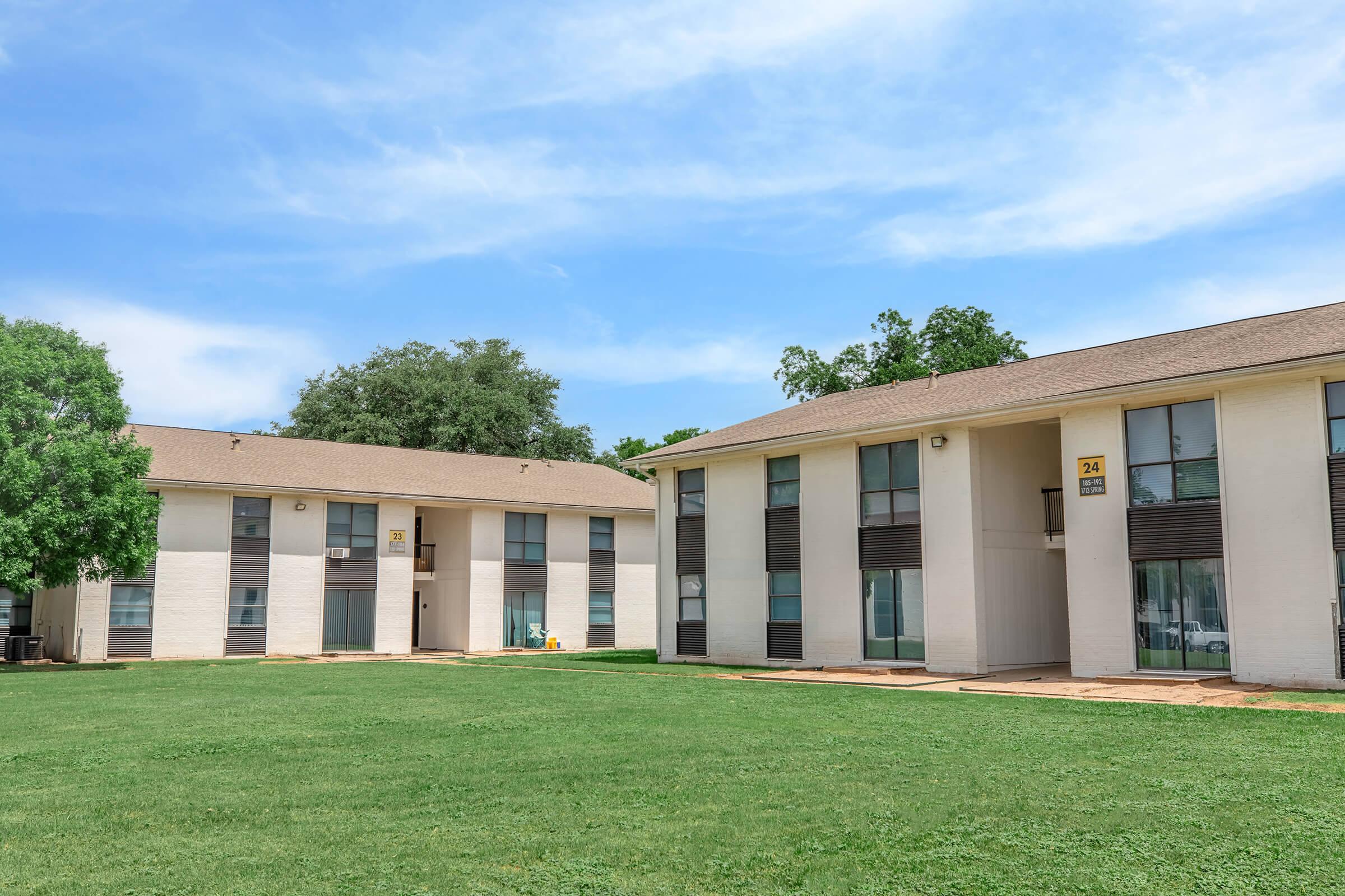 Exterior view of two multi-story residential buildings with several windows, surrounded by green grass and trees. The sky is clear with a few clouds. The buildings have a simple, modern design, and there is a small patio area visible in front of one of them.