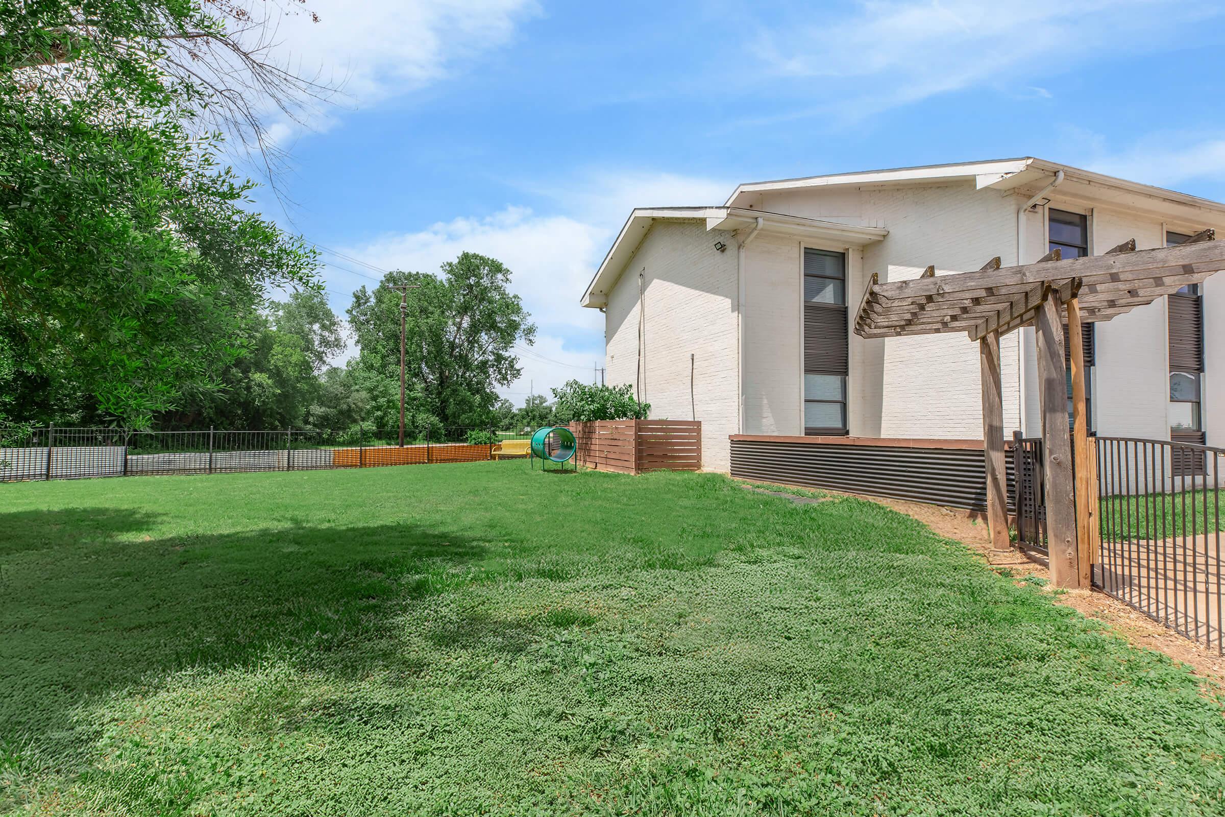 A well-maintained backyard of a residential property featuring green grass, a wooden pergola, and a fenced area. Lush trees provide shade, and a circular play structure can be seen in the distance. The house has a modern architectural style with large windows and light-colored siding.