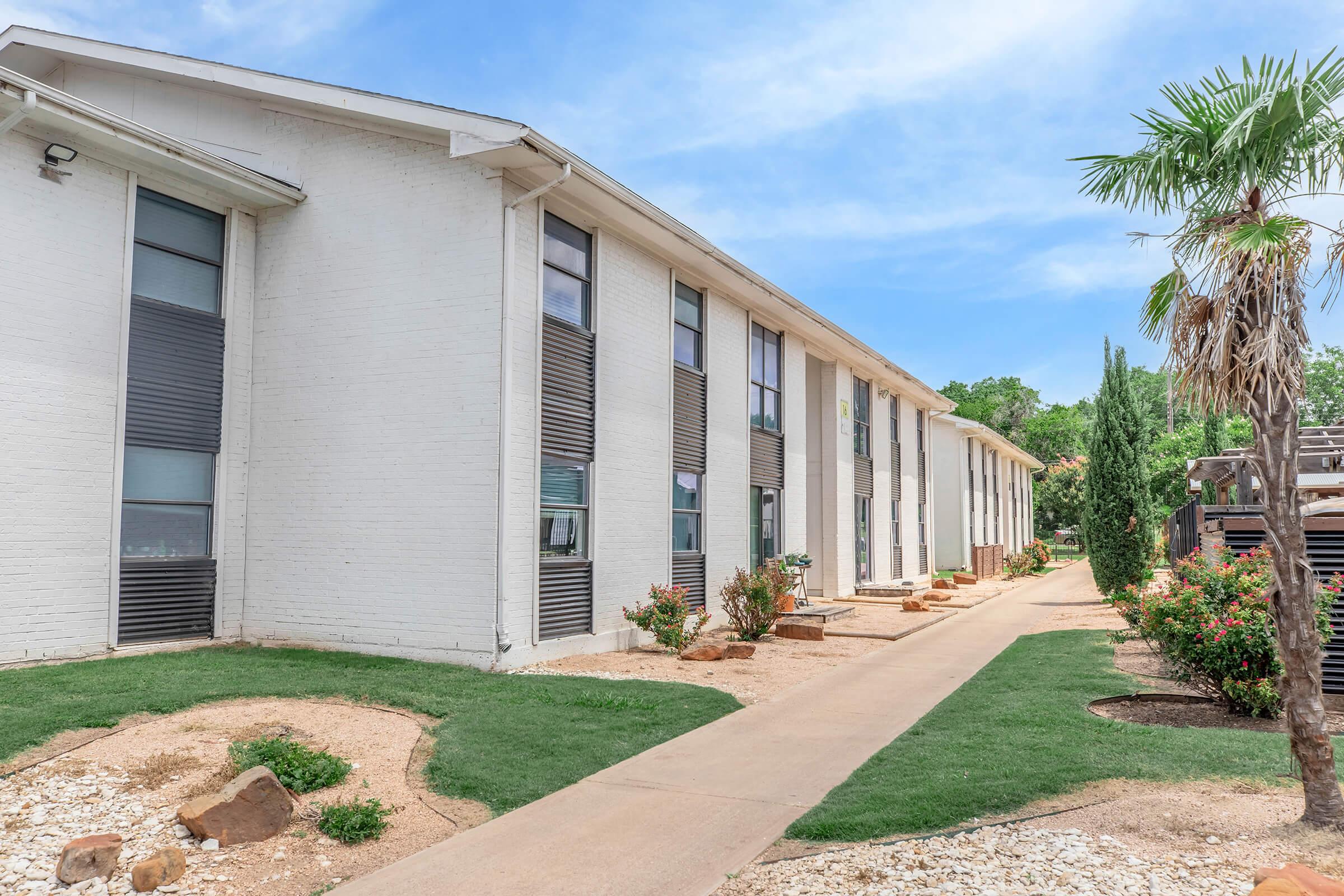 A modern, two-story building with a white exterior and large windows is seen along a paved pathway. The landscape includes neatly trimmed grass, decorative rocks, and shrubs. In the foreground, there are palm trees, and the sky is bright with a few clouds, indicating a sunny day.