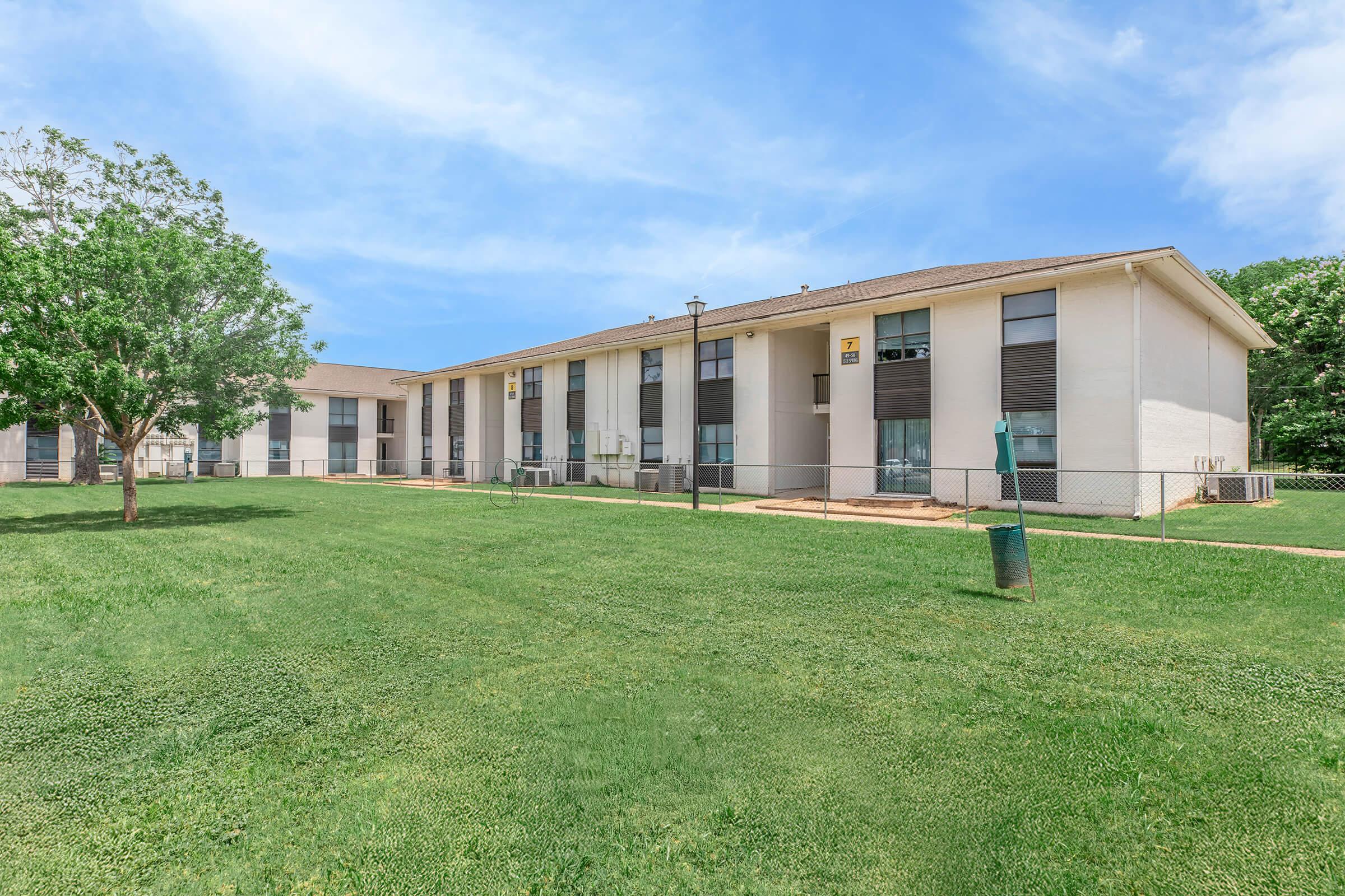 A residential apartment complex featuring two-story buildings with multiple windows. A well-maintained grassy area surrounds the buildings, and a tree is visible on the left. The sky is clear and blue, creating a pleasant setting.