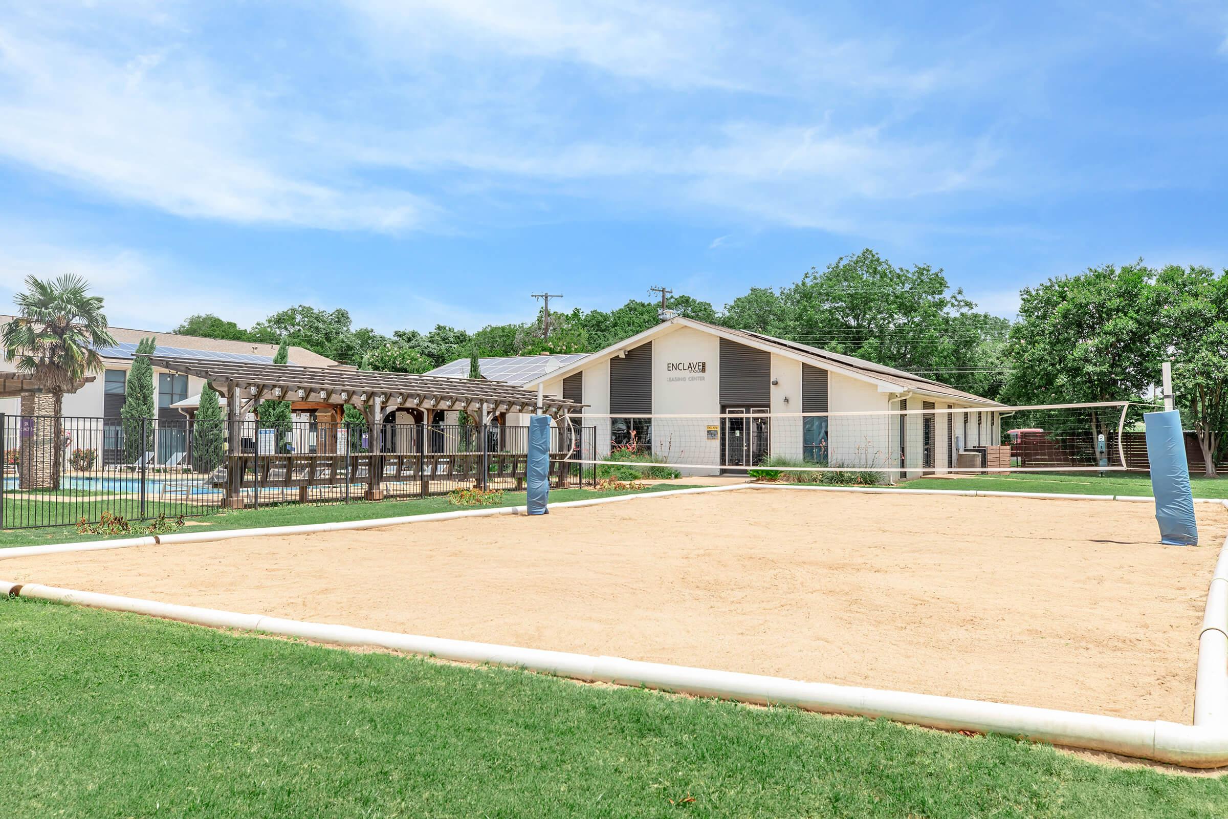A sandy volleyball court surrounded by green grass, with a fenced area for seating and leisure. In the background, there is a modern building with a sign, set amongst trees under a clear blue sky.
