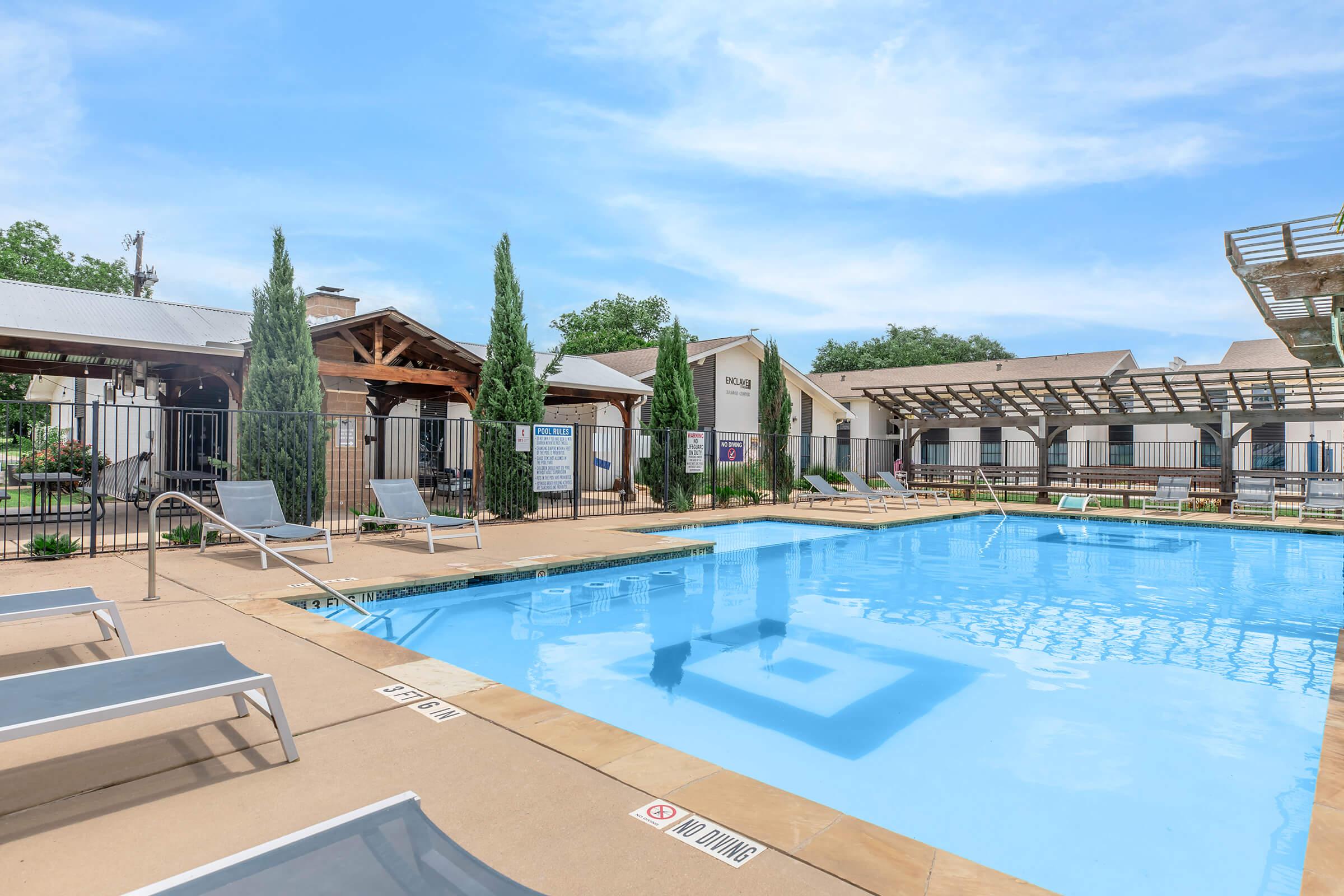 A clear swimming pool surrounded by lounge chairs, with green shrubs and trees nearby. In the background, there are wooden structures and a building. The sky is bright and blue, creating a sunny atmosphere at a recreational area. Signs and amenities are visible around the pool area.