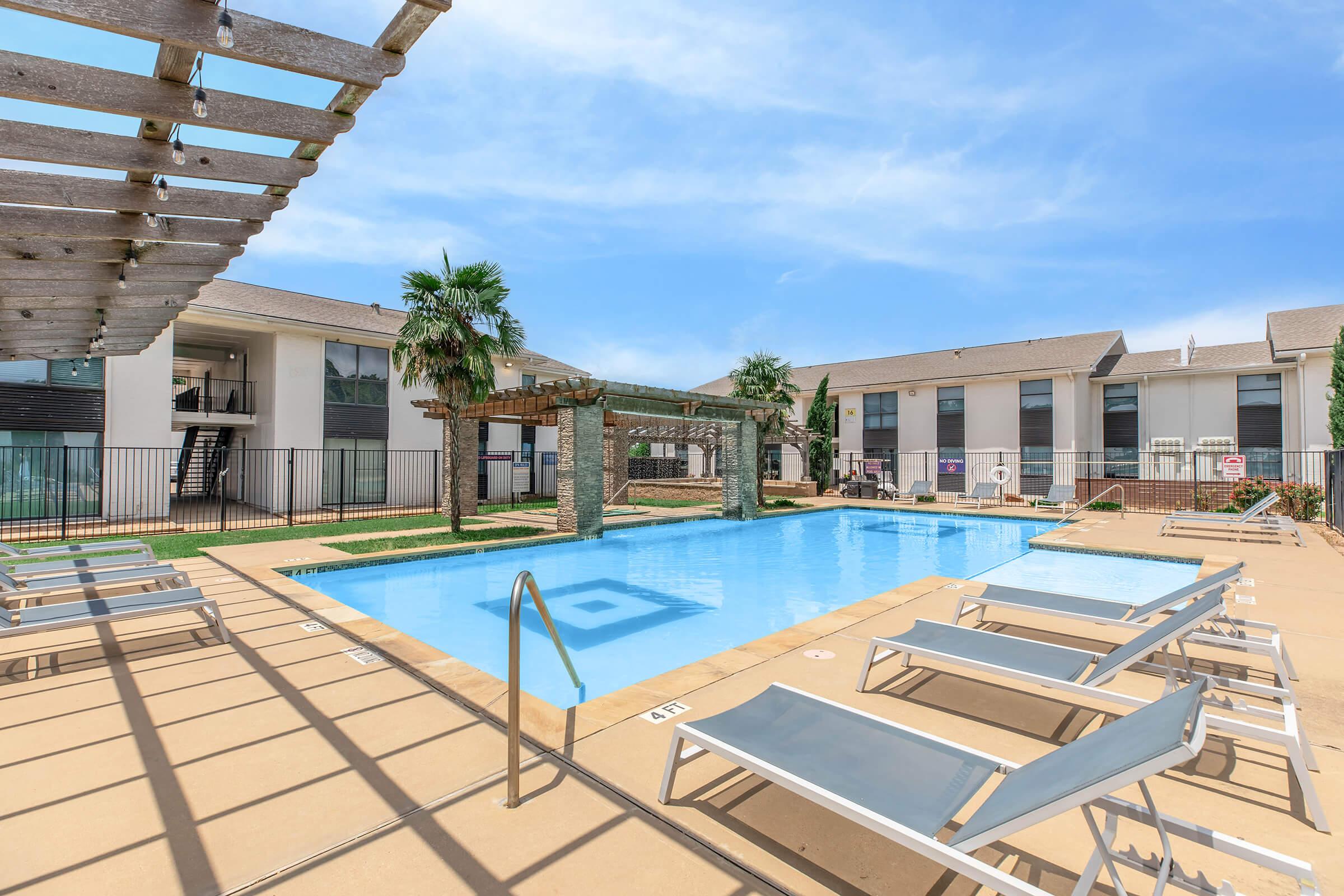 A clear blue swimming pool surrounded by lounge chairs and shaded areas, with a building complex in the background. Palm trees and a decorative pergola enhance the relaxing atmosphere of the outdoor space, ideal for leisure and recreation. Sunny weather adds to the inviting ambiance.