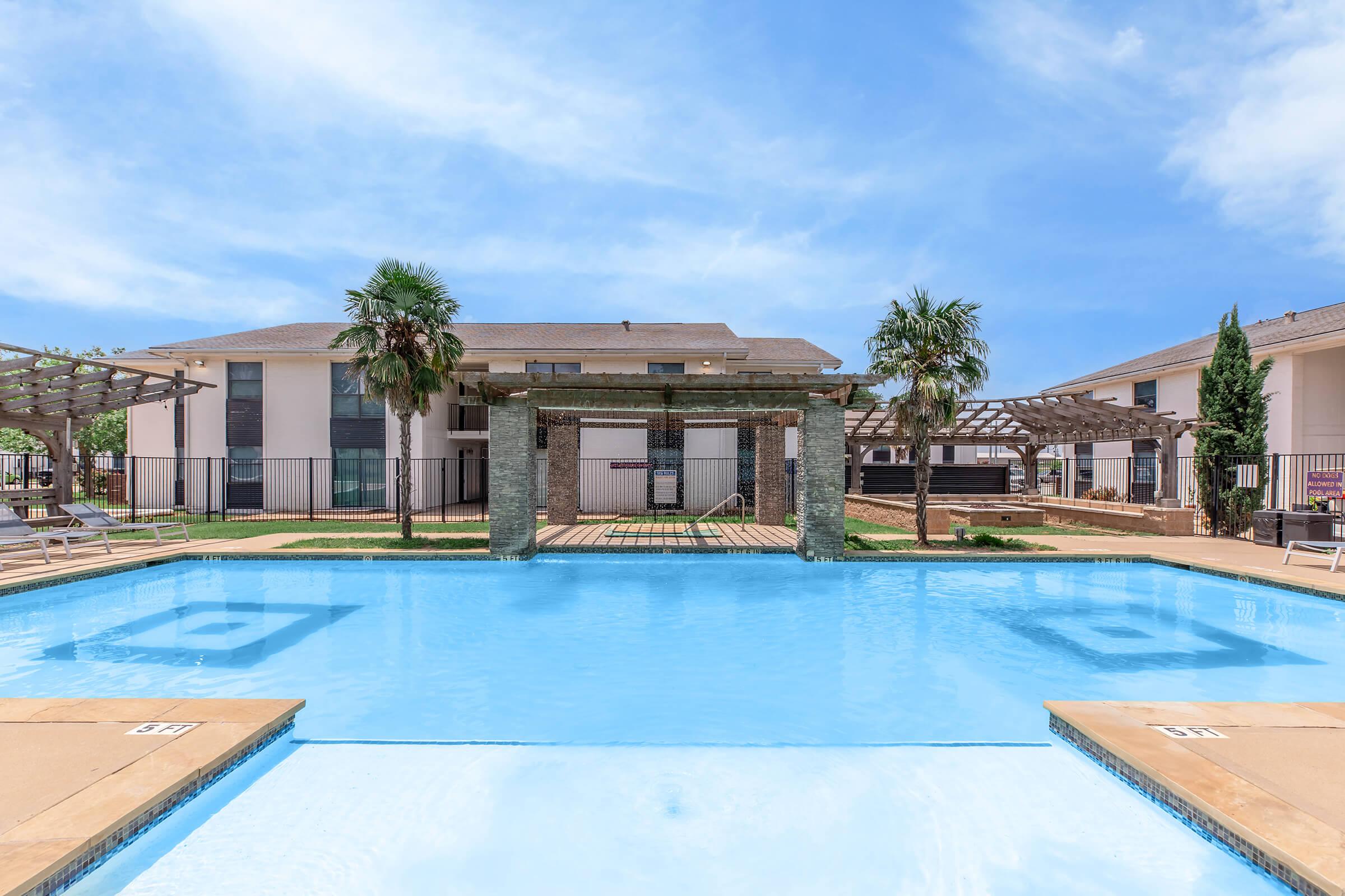 A clear blue swimming pool in the foreground with lounge chairs surrounding it. In the background, there are two apartment buildings, one with a pergola and palm trees. The sky is bright and clear, contributing to a sunny outdoor atmosphere.