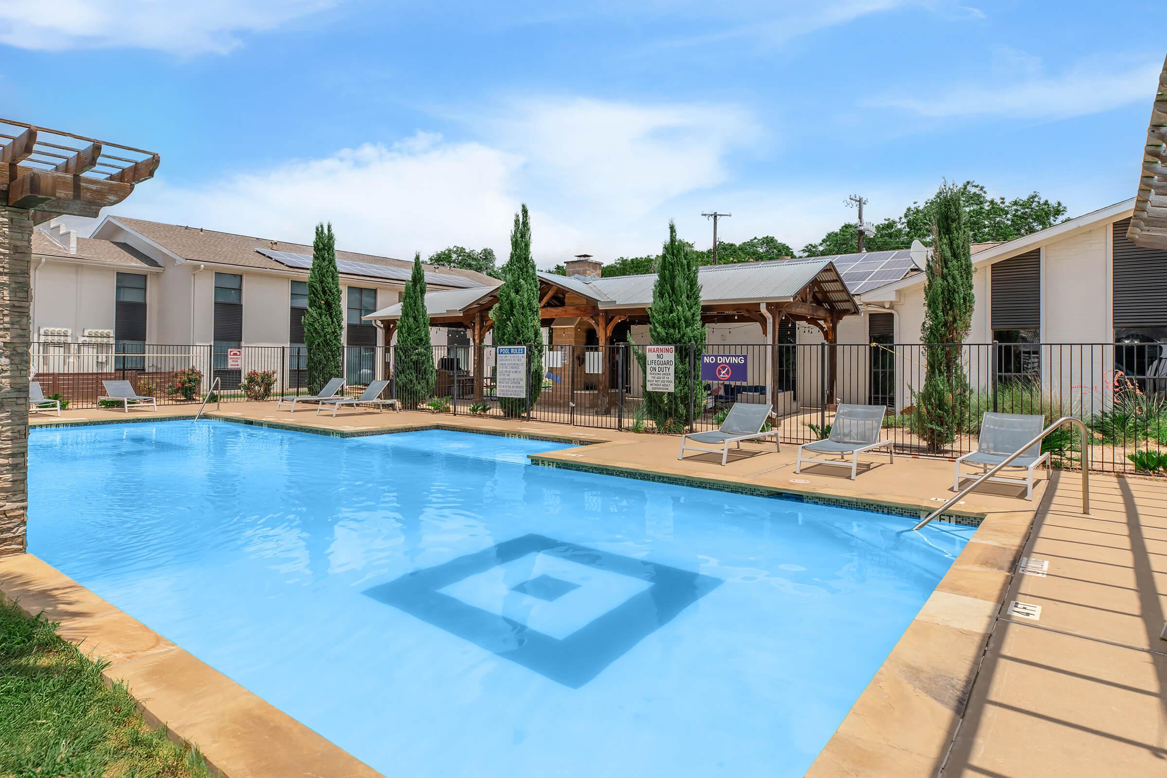 A serene outdoor swimming pool area featuring clear blue water, surrounded by lounge chairs, tall greenery, and shaded structures. The pool is enclosed with a fence, and several signs are visible around the area, enhancing the inviting atmosphere for relaxation and leisure.