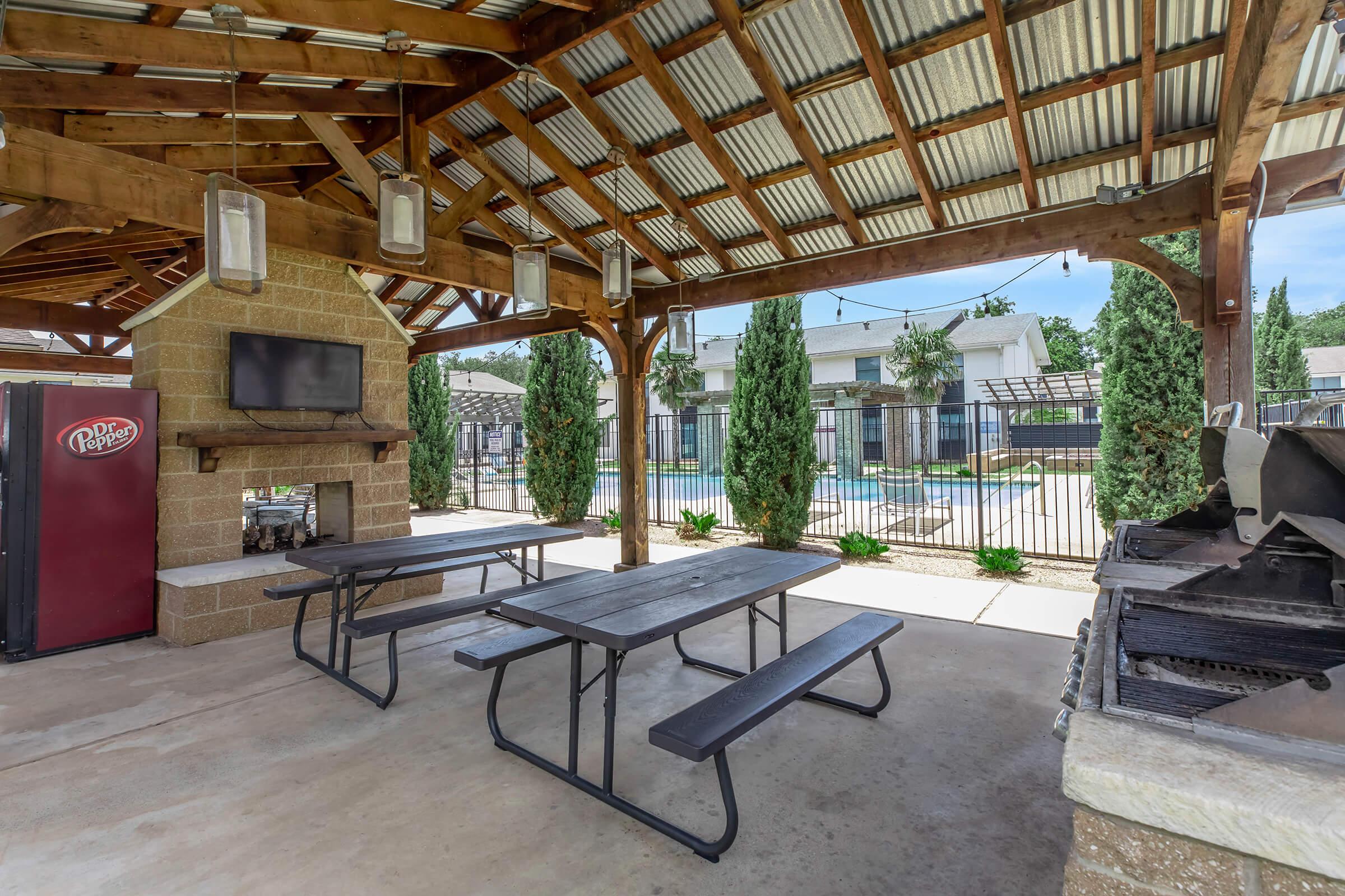 Outdoor pavilion with a wooden roof, featuring a picnic area with black tables and benches. There's a stone fireplace, a television mounted on the wall, and a view of a swimming pool surrounded by greenery. The area is neatly arranged, ideal for gatherings and relaxation.