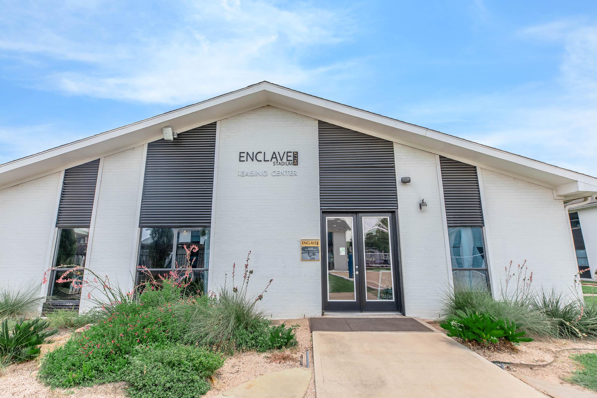 Leasing center building with modern architecture featuring large front windows and a sloped roof. The name "Enclave" is displayed prominently above the entrance. Surrounding the entrance are landscaped areas with plants and gravel. The overall design is contemporary and welcoming.