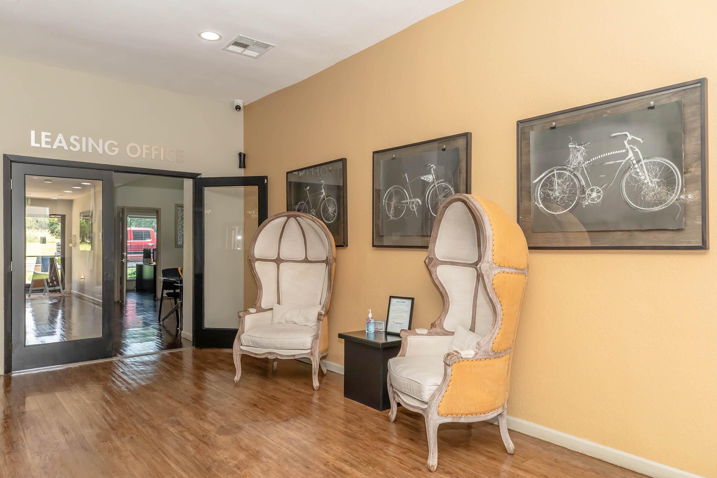 Interior of a leasing office featuring two stylish high-backed chairs in cream and tan, a dark side table with a paper on it, and three framed black-and-white bicycle prints on the wall. The walls are painted a warm tan color, and glass doors lead to another room in the background. Brightly lit and welcoming atmosphere.