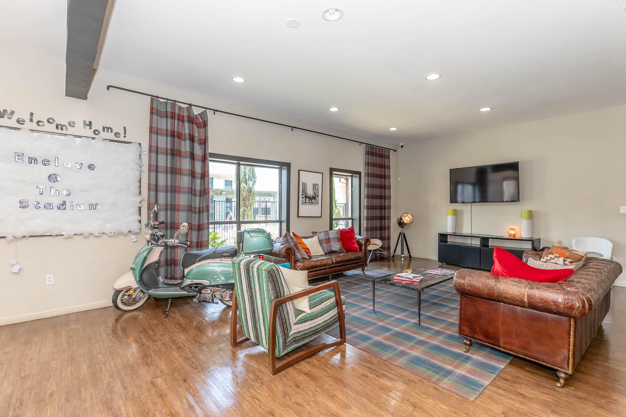 A cozy living room with hardwood floors, featuring a leather couch, modern chairs, and a green vintage scooter. Brightly colored pillows add a pop of color. A large TV is mounted on the wall, and curtains in plaid pattern frame the windows. Decorative elements like a welcome sign and a small lamp enhance the inviting atmosphere.