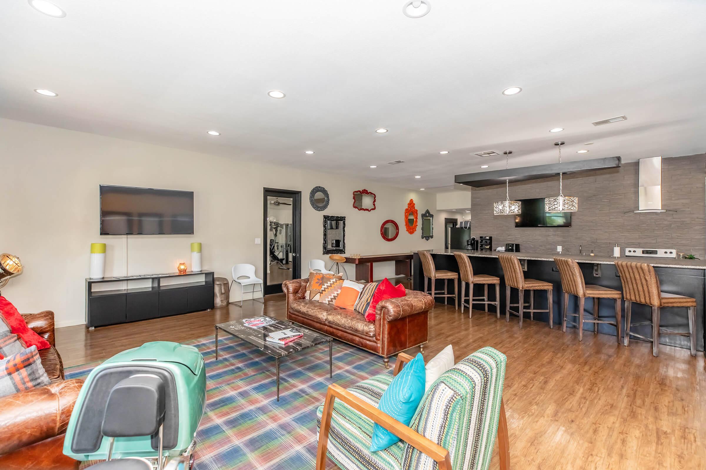 Modern living room and kitchen area with hardwood floors. The space features a leather sofa, a colorful armchair, and a coffee table on a patterned rug. A bar with high stools separates the kitchen, equipped with sleek appliances and a flat-screen TV mounted on the wall. Bright lighting enhances the open layout.