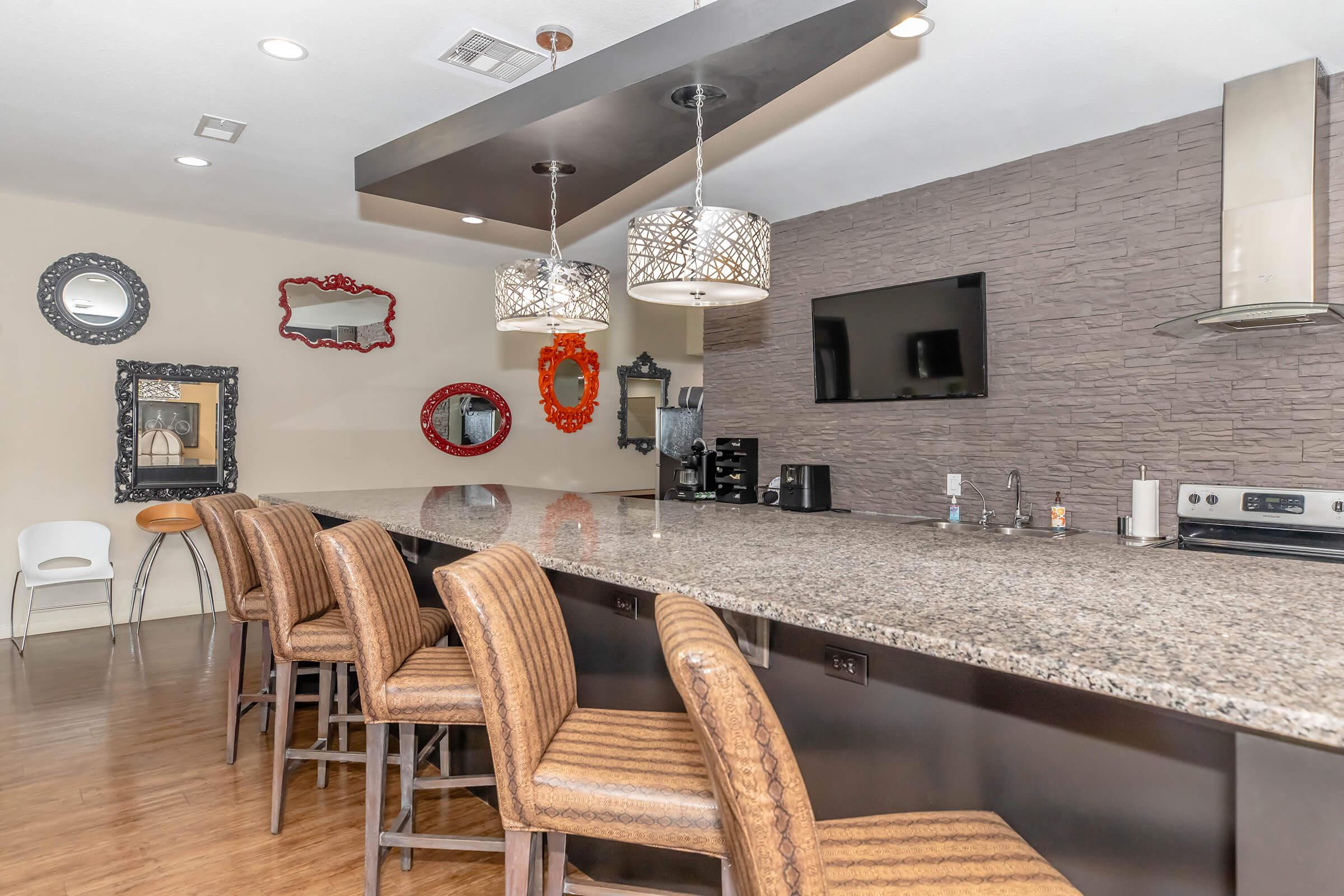 A modern kitchen with a sleek, dark granite countertop and stylish bar stools. Decorative mirrors in various shapes and colors hang on the wall, and pendant lights illuminate the space. A wall-mounted TV and kitchen appliances are also visible, creating a contemporary and inviting atmosphere.