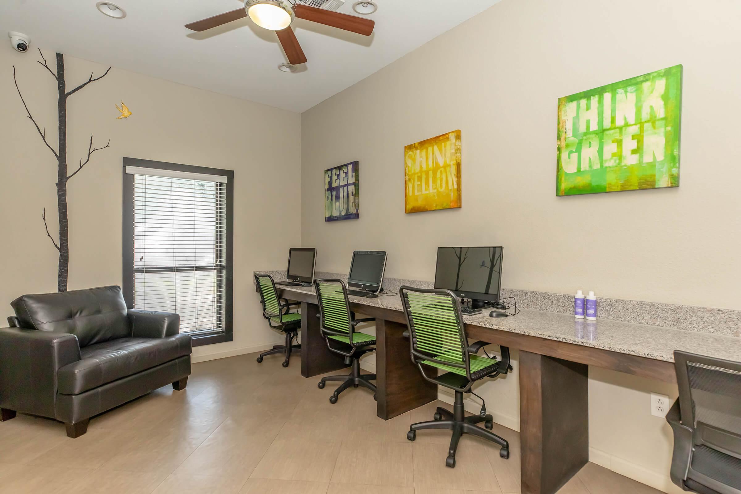 A workspace featuring two computer desks with green office chairs, a black leather armchair, and colorful motivational wall art that reads "FEEL MUCH," "SHINE YELLOW," and "THINK GREEN." Natural light comes through the window, and a simple tree mural is painted on the wall.