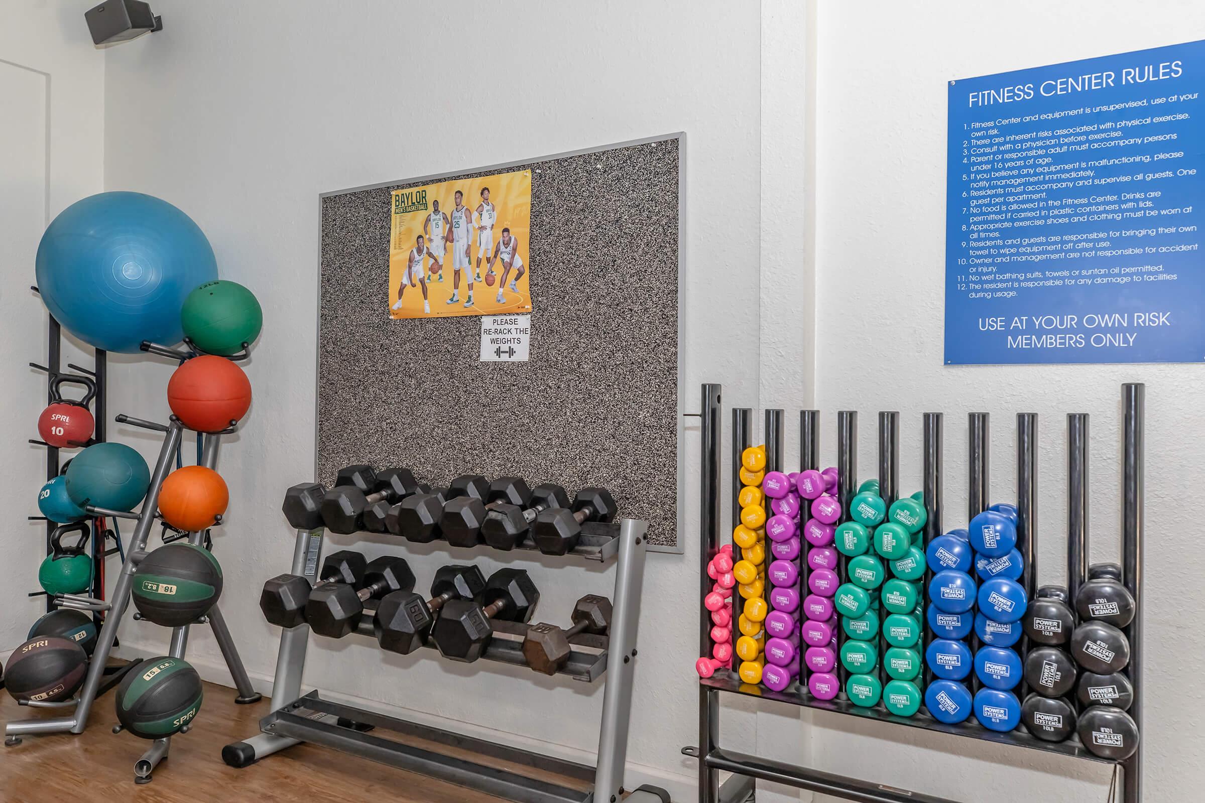 A fitness area featuring colorful dumbbells arranged on a rack, various exercise balls, and a bulletin board with fitness posters. A sign detailing fitness center rules is mounted on the wall, emphasizing safety and membership guidelines.