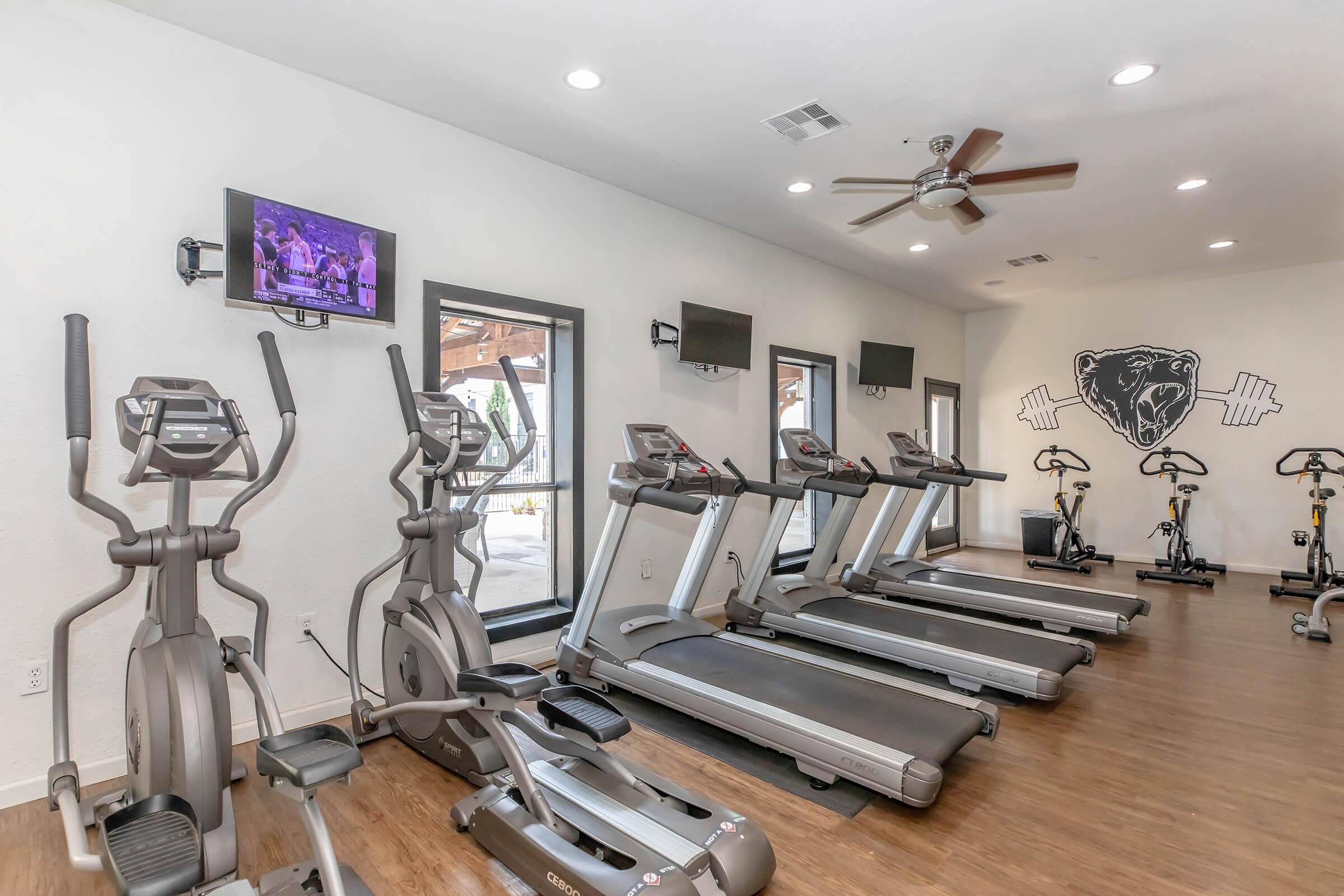 A spacious gym area featuring several treadmills and ellipticals, equipped with multiple wall-mounted TVs for entertainment. Large windows provide natural light, and a ceiling fan adds ventilation to the workout space.