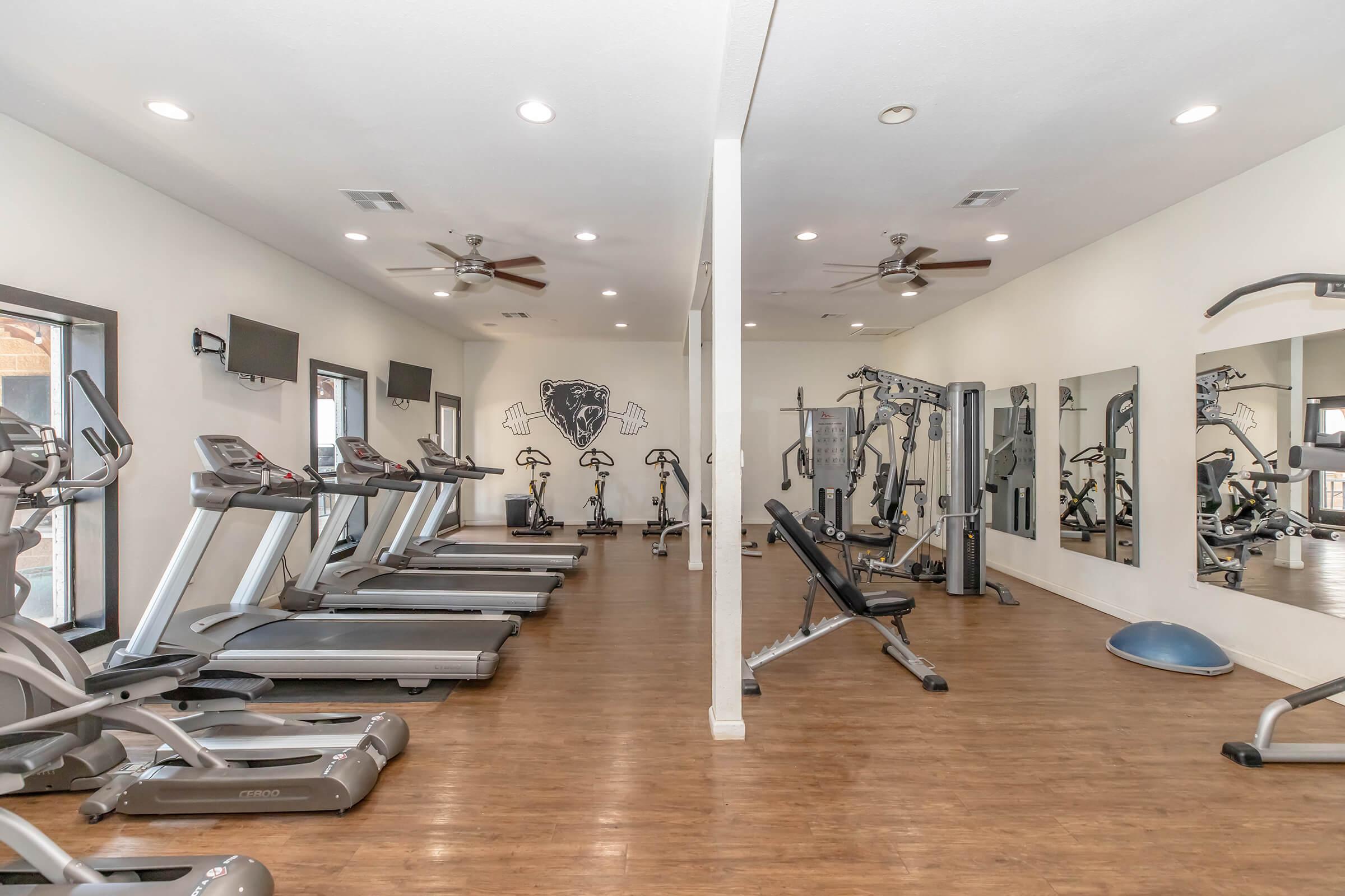 A spacious gym featuring several treadmills, weight machines, and free weights. The room has a modern design with large mirrors on the wall, ceiling fans, and multiple flat-screen TVs. Natural light enters through large windows, enhancing the open and inviting atmosphere.