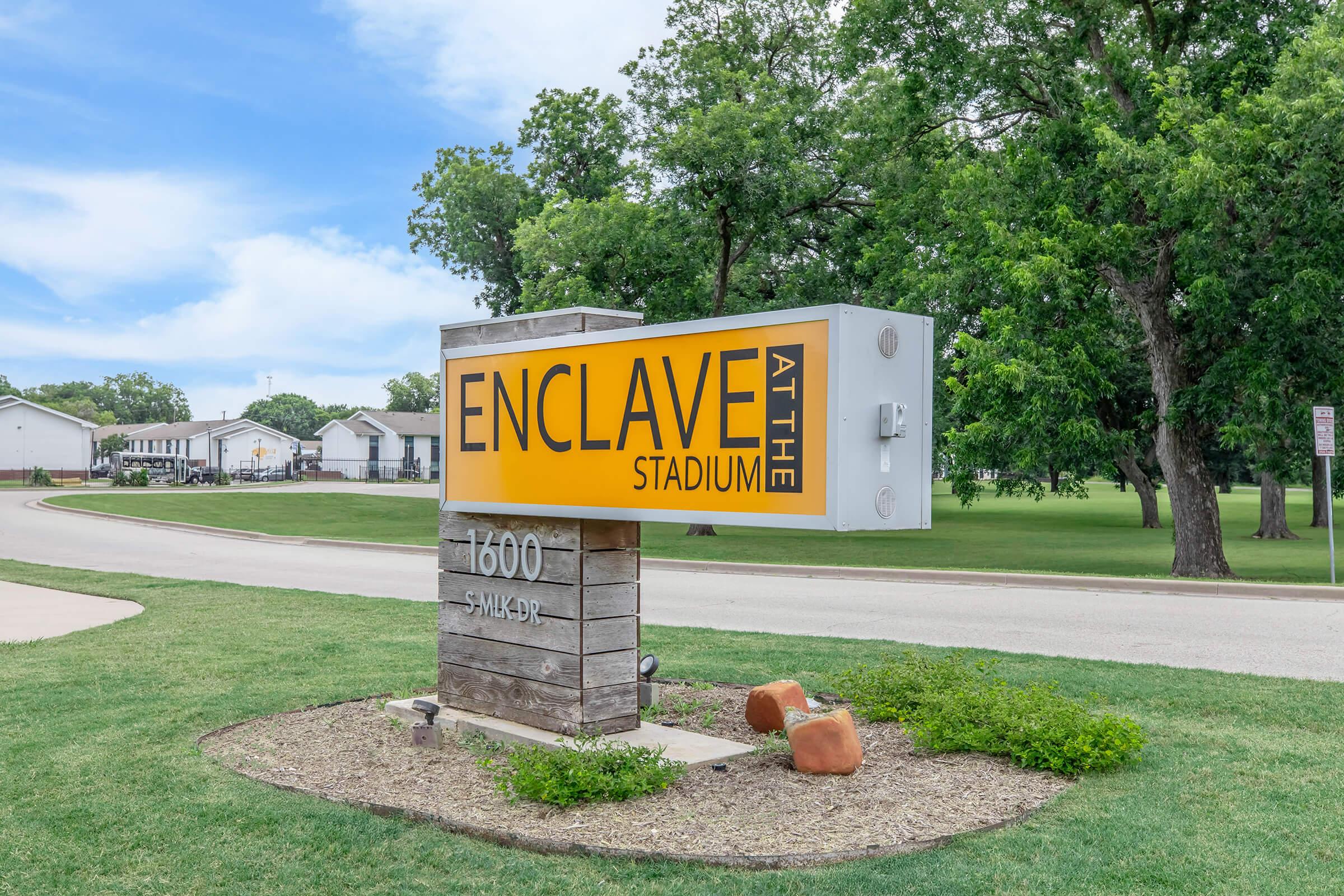 Sign for "Enclave at the Stadium" featuring a prominent yellow background with black text. The address "1600 S. May" is displayed below. The sign is surrounded by green grass and trees, with residential buildings visible in the background.