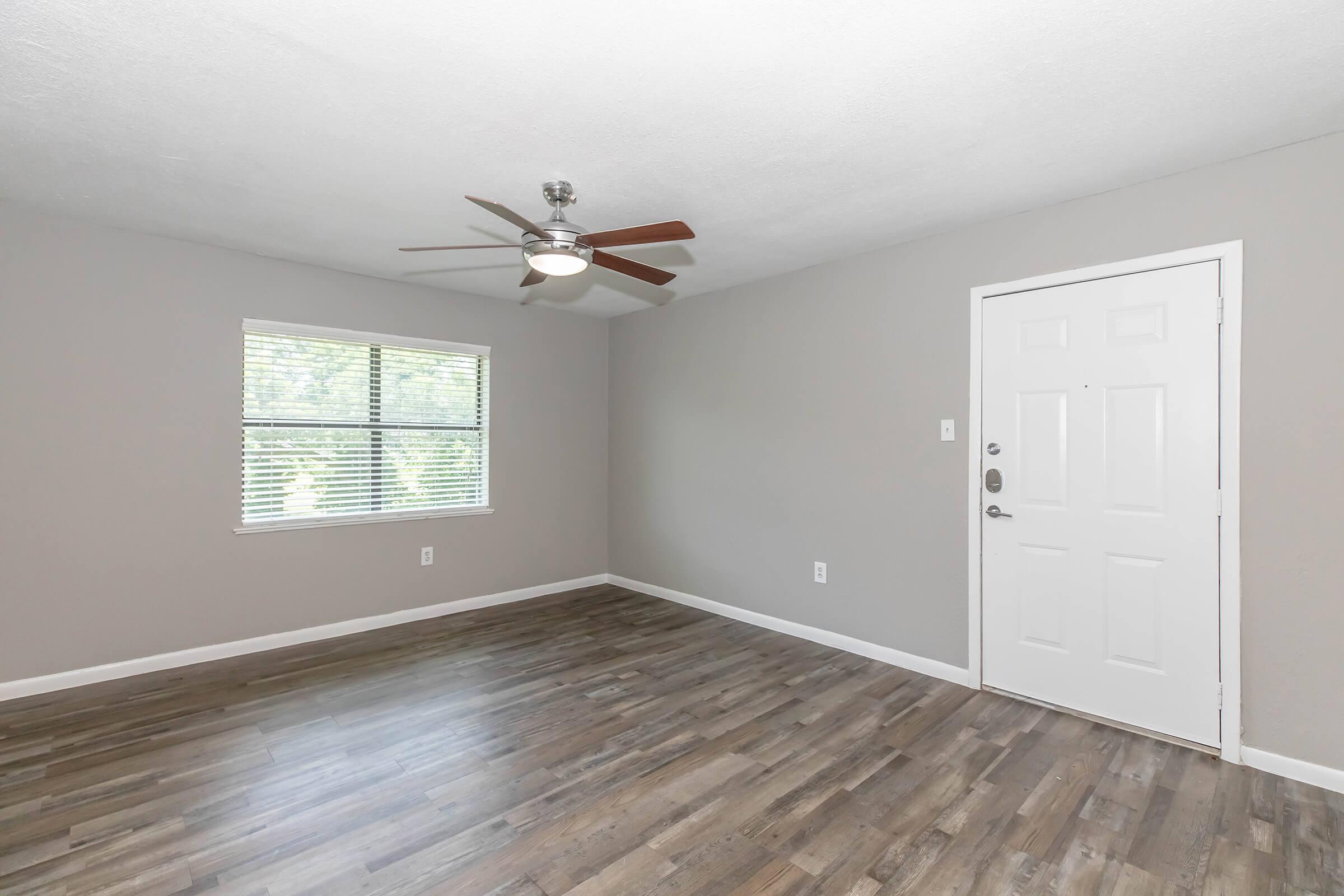 An empty room with light gray walls and a ceiling fan. A window with blinds allows natural light, and there is a front door on the right. The floor is covered in wooden laminate, creating a clean and modern look.