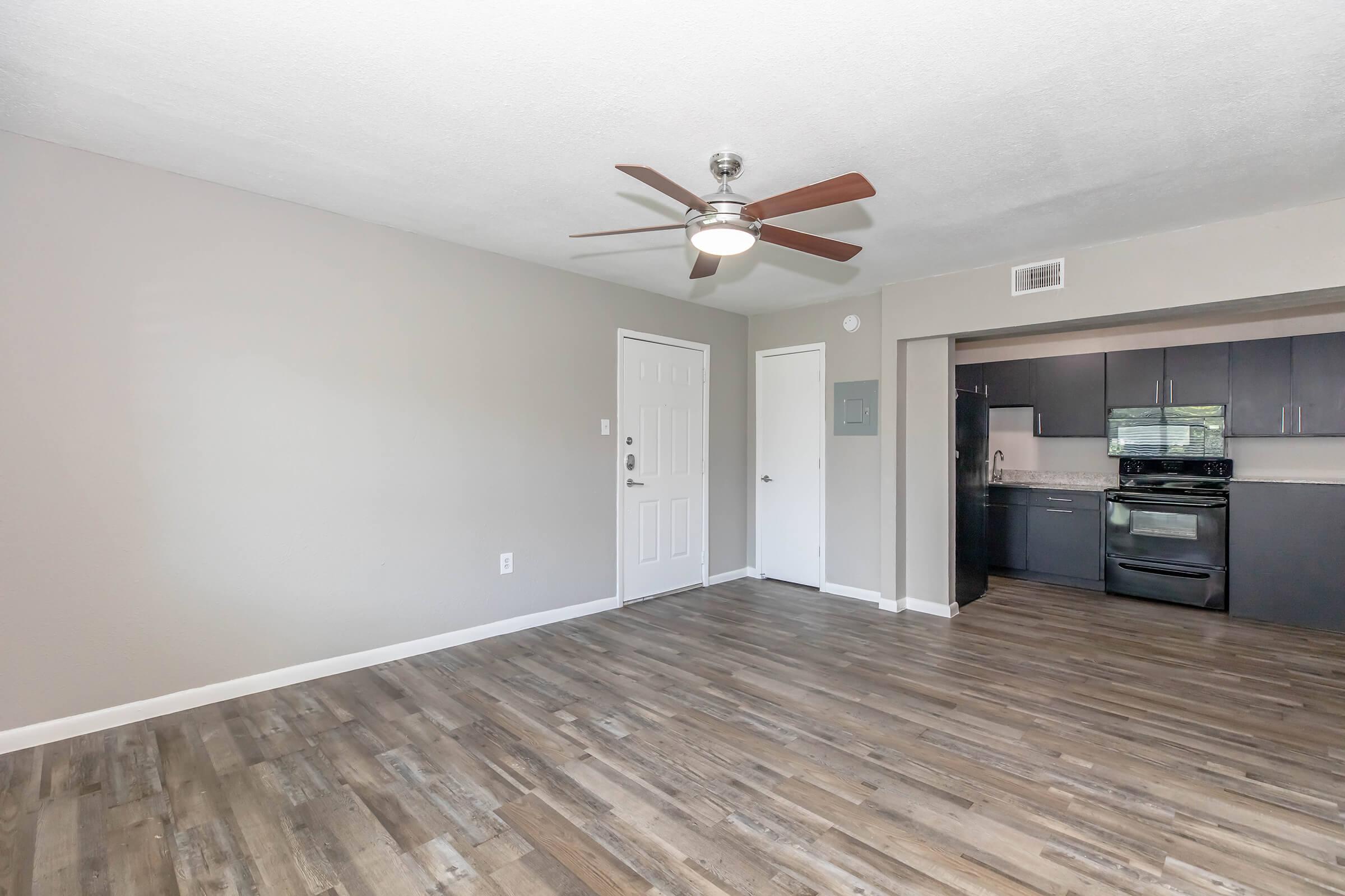 Spacious living area with light-colored walls and modern ceiling fan, leading to a door. A stylish kitchen with dark cabinetry and stainless steel appliances is visible in the background, along with wood-look flooring throughout the space.