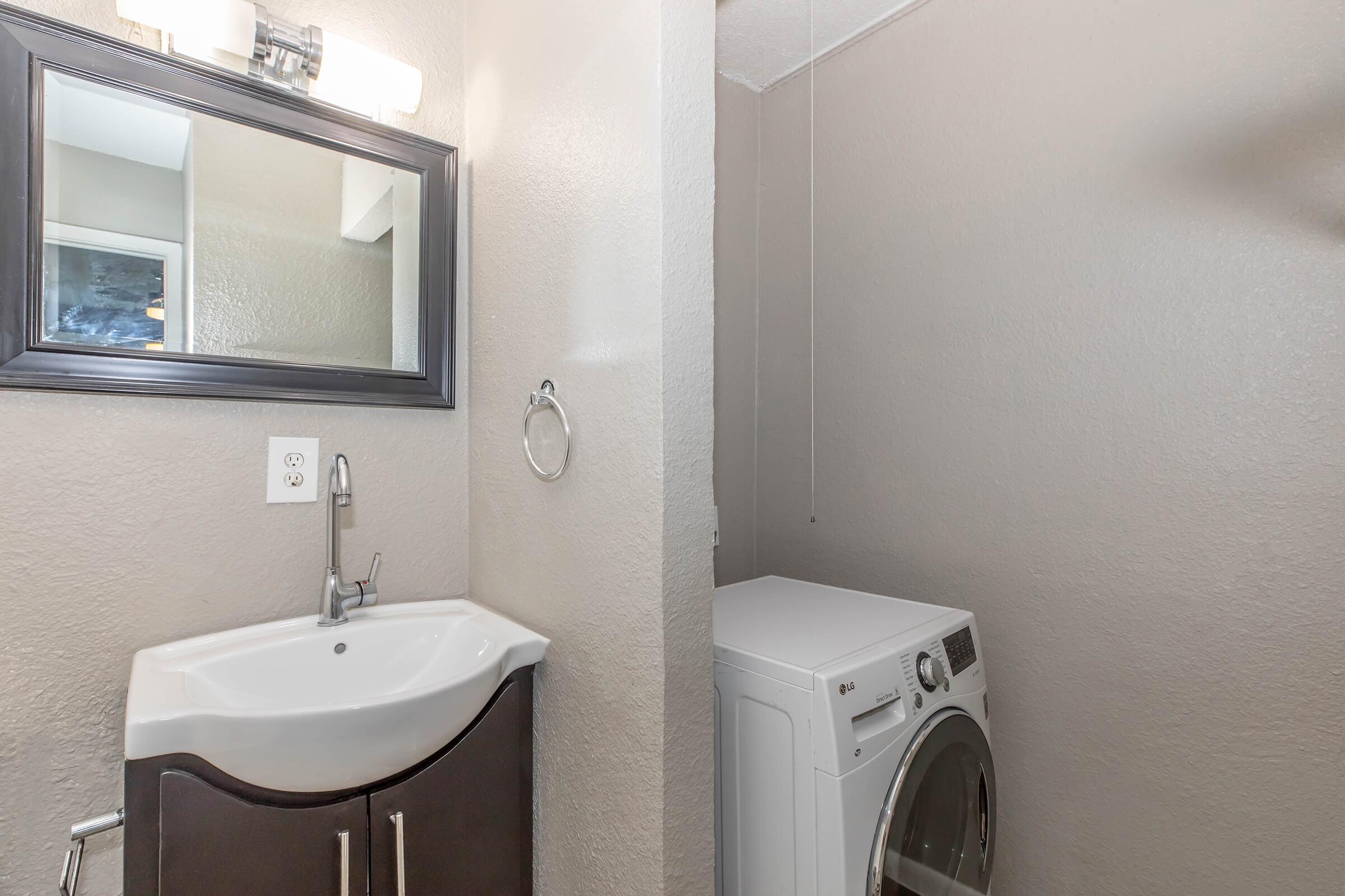 A small laundry area featuring a modern washing machine beside a compact sink with a stylish faucet. The walls are painted in a light color, and thereโs a mirror above the sink. The space is well-lit, creating a clean and functional atmosphere.