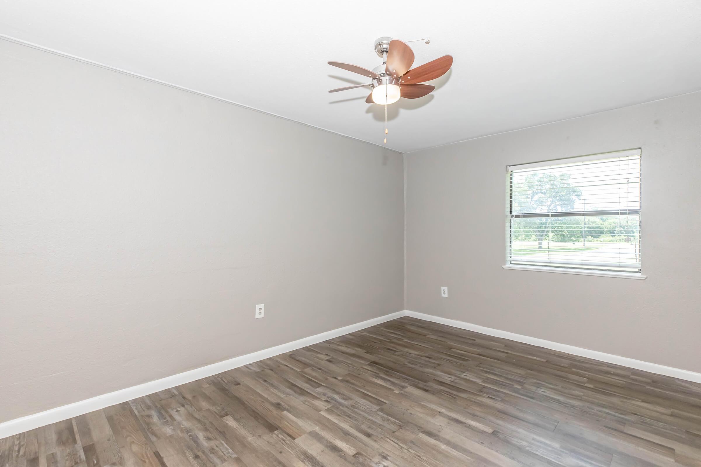 A simple, empty room with light brown walls and a ceiling fan featuring wooden blades. The floor is a medium-toned laminate wood, and there is a window with blinds allowing natural light to enter the space.