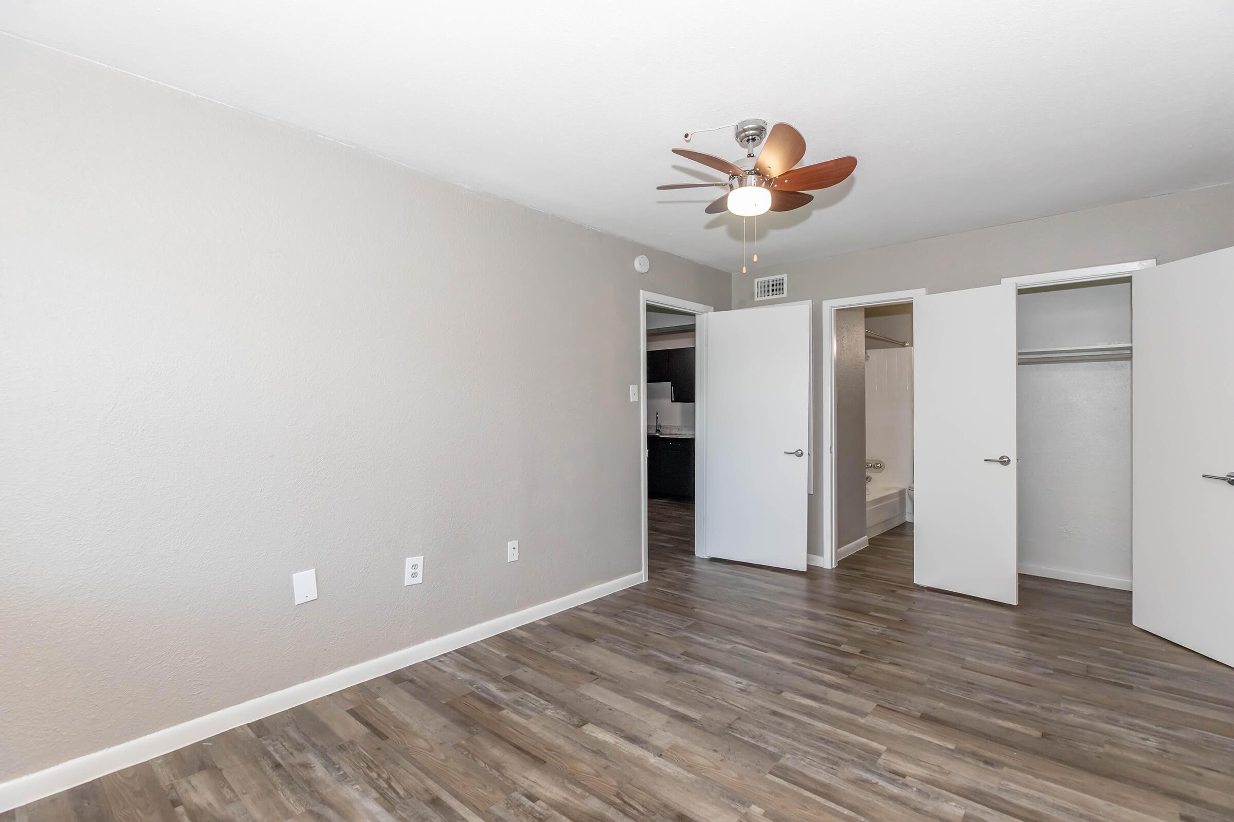 A spacious, well-lit room featuring light gray walls, a ceiling fan with wooden blades, and laminate flooring. Two white doors lead to a closet and bathroom, while the opposite wall remains empty, creating an inviting and open atmosphere.