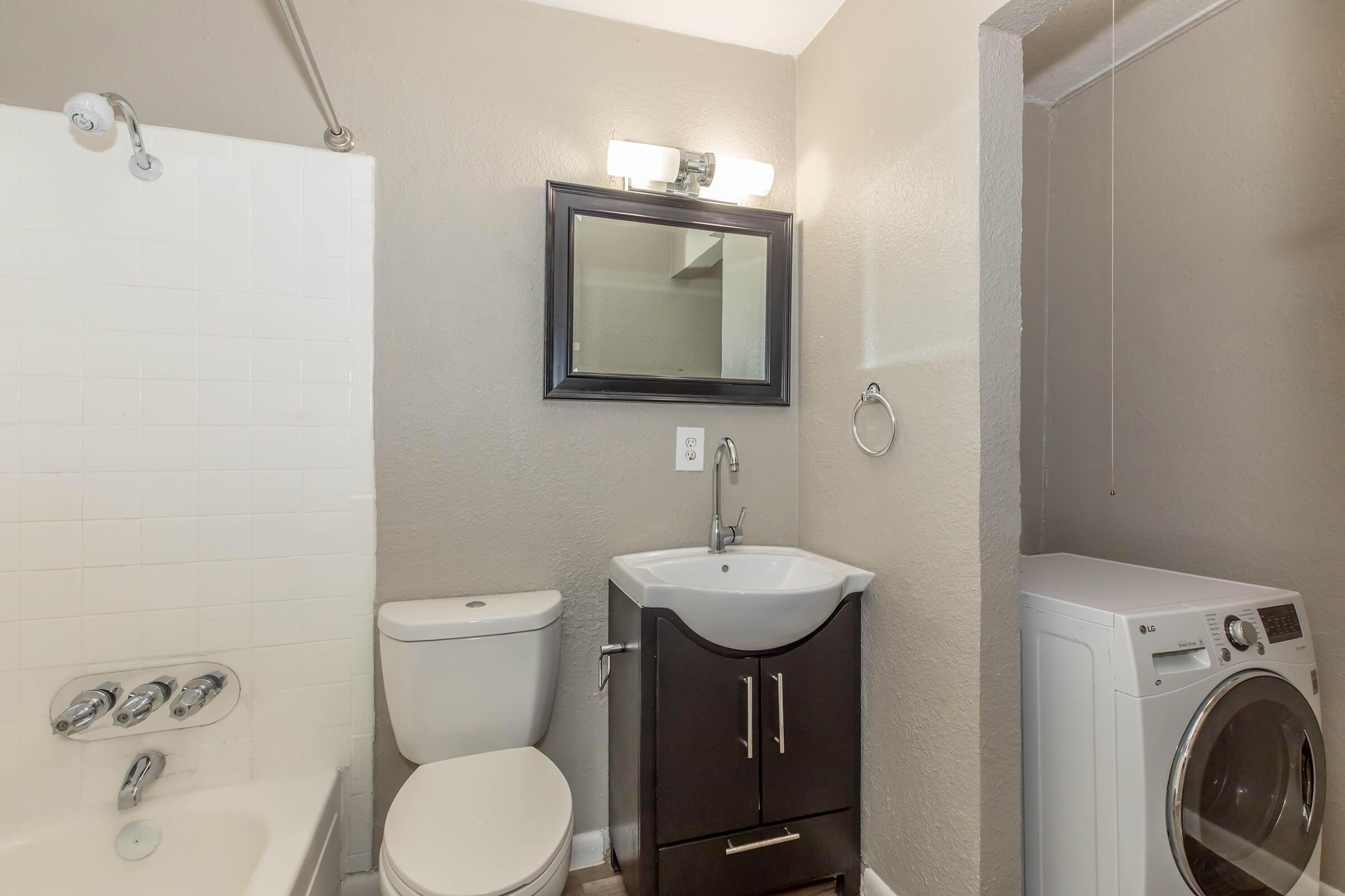 A small, modern bathroom featuring a white bathtub with a shower, a dark wood vanity with a sink, a toilet, and a washing machine in the corner. The walls are painted in a light gray color, and there is a rectangular mirror above the sink with a light fixture above it.