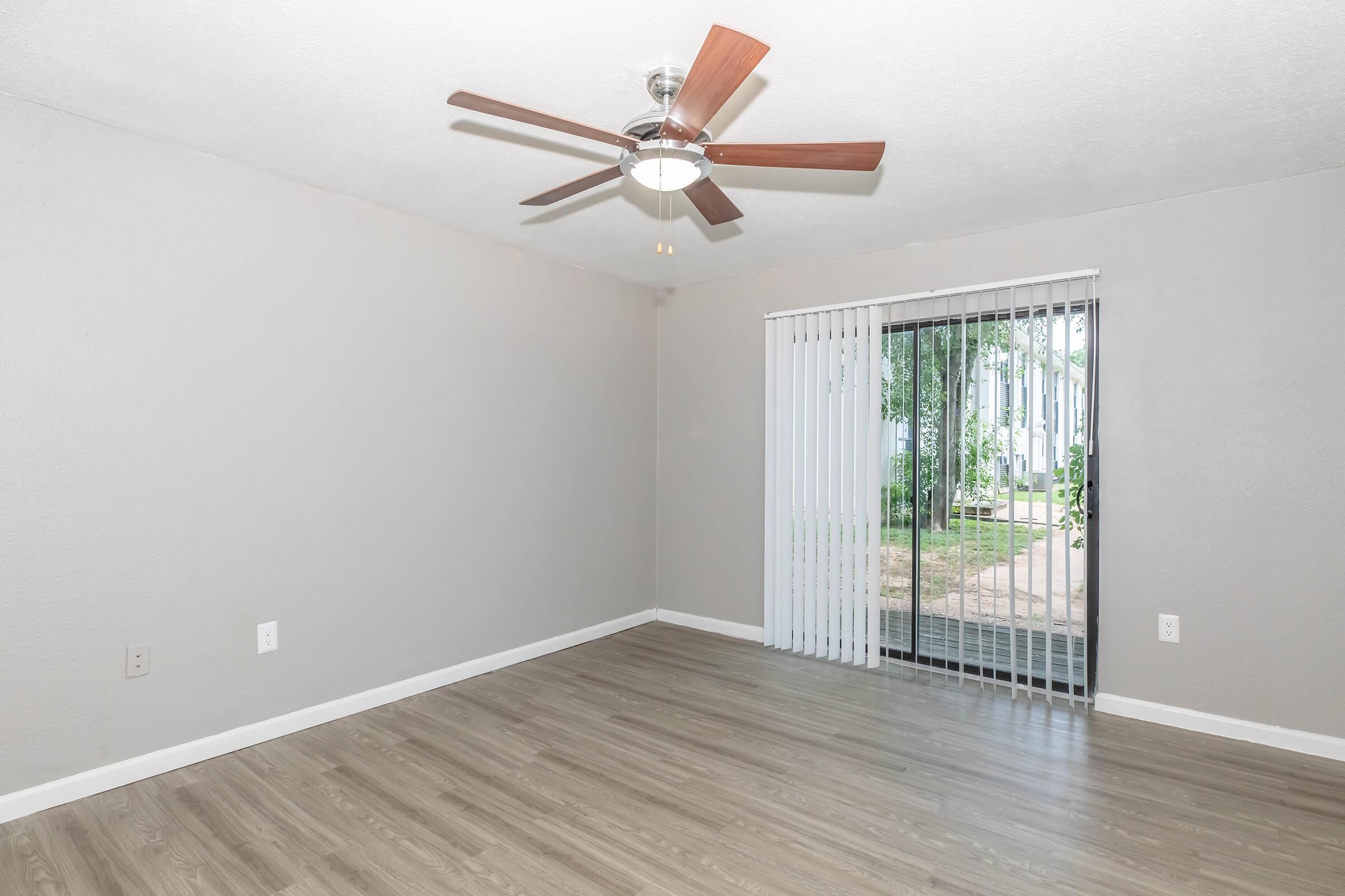 A spacious, empty room featuring light gray walls, a ceiling fan with wooden blades, and large sliding glass doors that lead to an outdoor area. The floor is a light wood laminate, creating a modern and inviting atmosphere. Natural light comes in through the blinds.