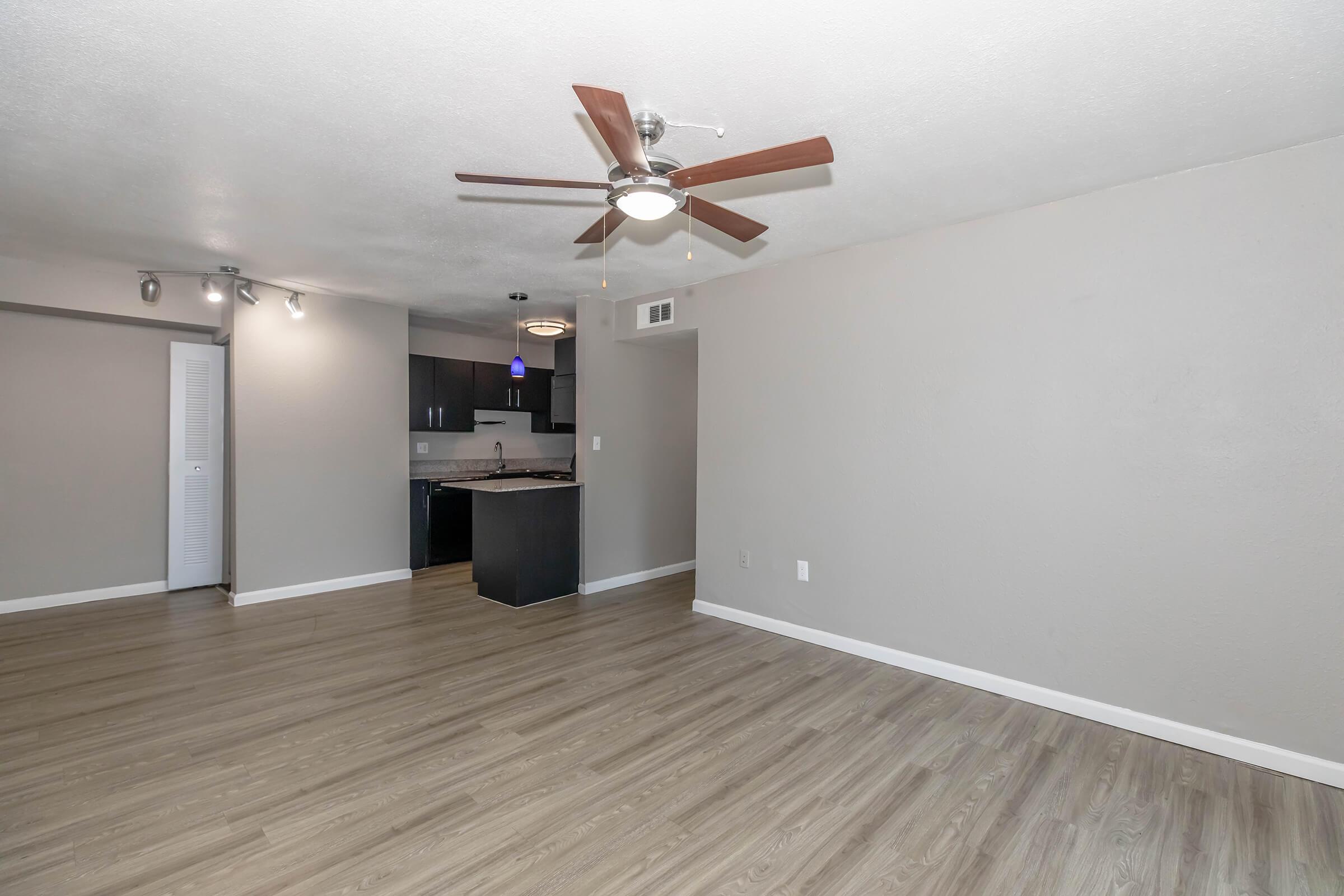 A spacious living area featuring light wood flooring, a ceiling fan with wooden blades, and neutral-colored walls. On one side, there is a wall-mounted light fixture and a view of an open kitchen with dark cabinets and a bar countertop. The space is well-lit, creating a modern and inviting atmosphere.