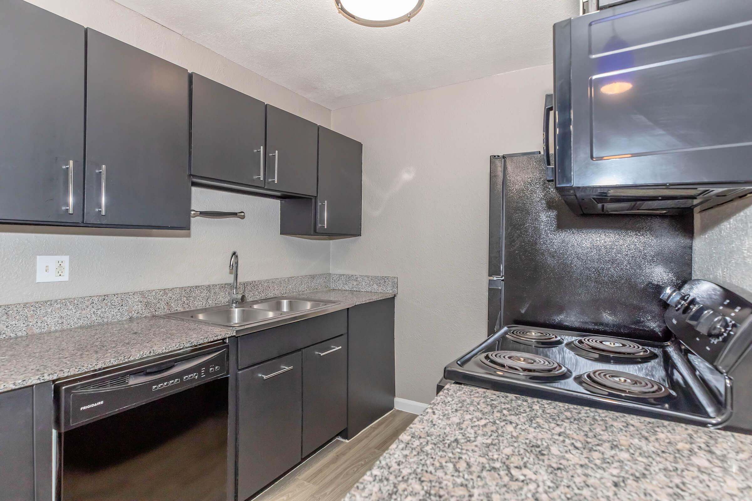 A modern kitchen featuring dark cabinets, a dual sink, a black refrigerator, and a stove with four burners. The countertop is made of granite, and the flooring is light-colored laminate, creating a bright and contemporary look.