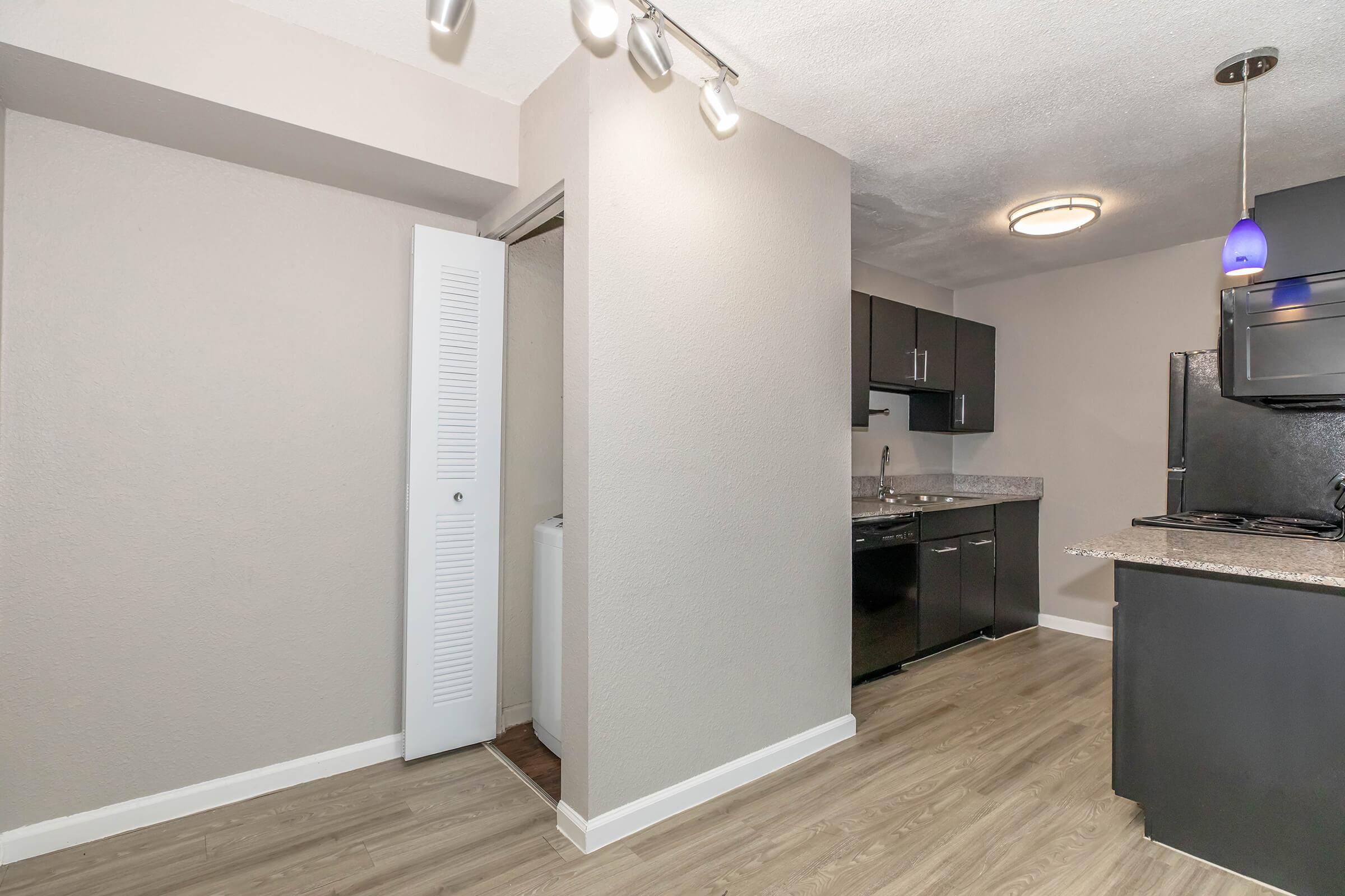 A modern kitchen space featuring dark cabinetry, a granite countertop, and stainless steel appliances. The area transitions into a small hallway with a white door leading to a laundry closet. Soft lighting is provided by an overhead fixture and a blue pendant light above the countertop. The floor is light-colored laminate.