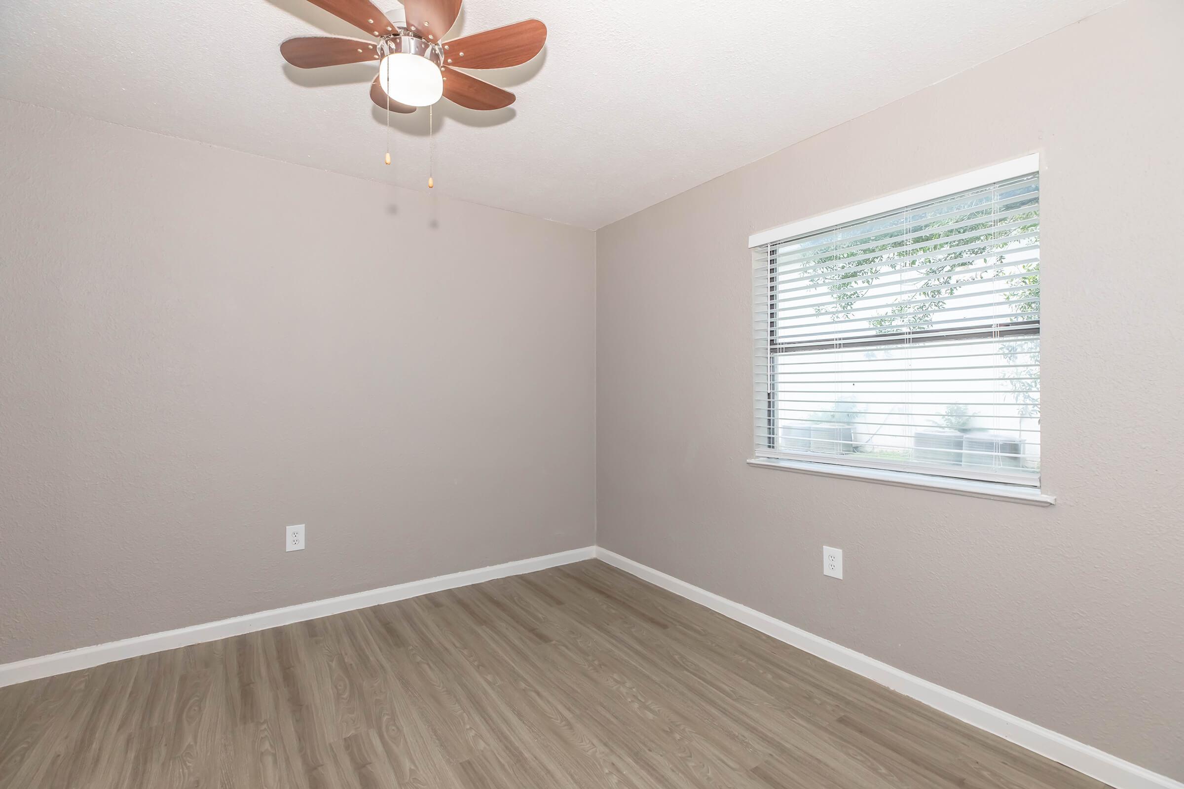 Empty room with light beige walls, a ceiling fan with wooden blades, and a window featuring white blinds. The floor is light wood laminate. There are no furnishings or decorations, creating a minimalistic and spacious atmosphere.