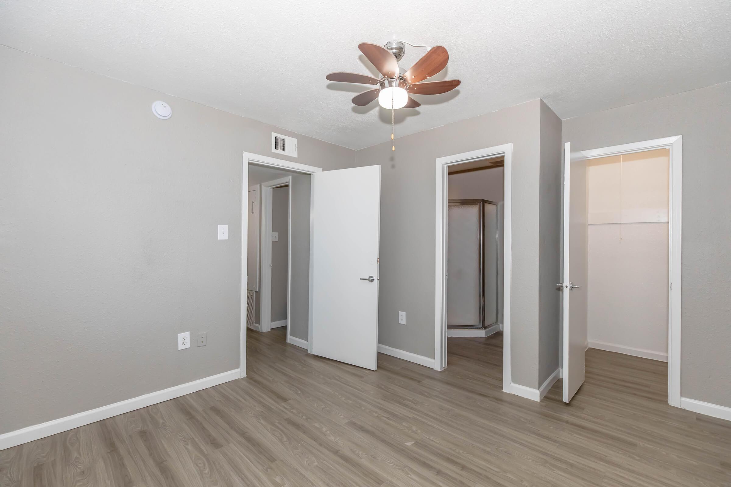 A neutral-colored room featuring a ceiling fan with wooden blades. There are two doorways leading to adjacent spaces, one with a sliding door. The floor is light-colored laminate, and the walls are painted in a soft gray. Natural light brightens the area, giving it a spacious feel.