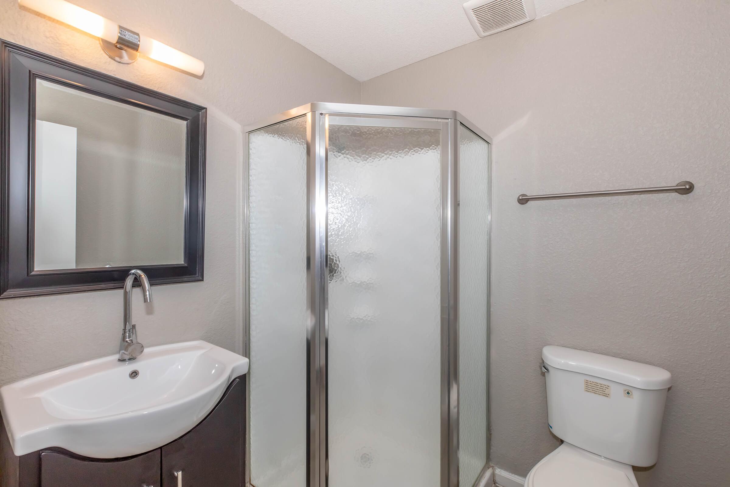 A modern bathroom featuring a shower with a glass enclosure, a sink with a curved basin, a dark-framed mirror above the sink, and a white toilet. The walls are painted in a soft gray color, creating a clean and contemporary atmosphere.